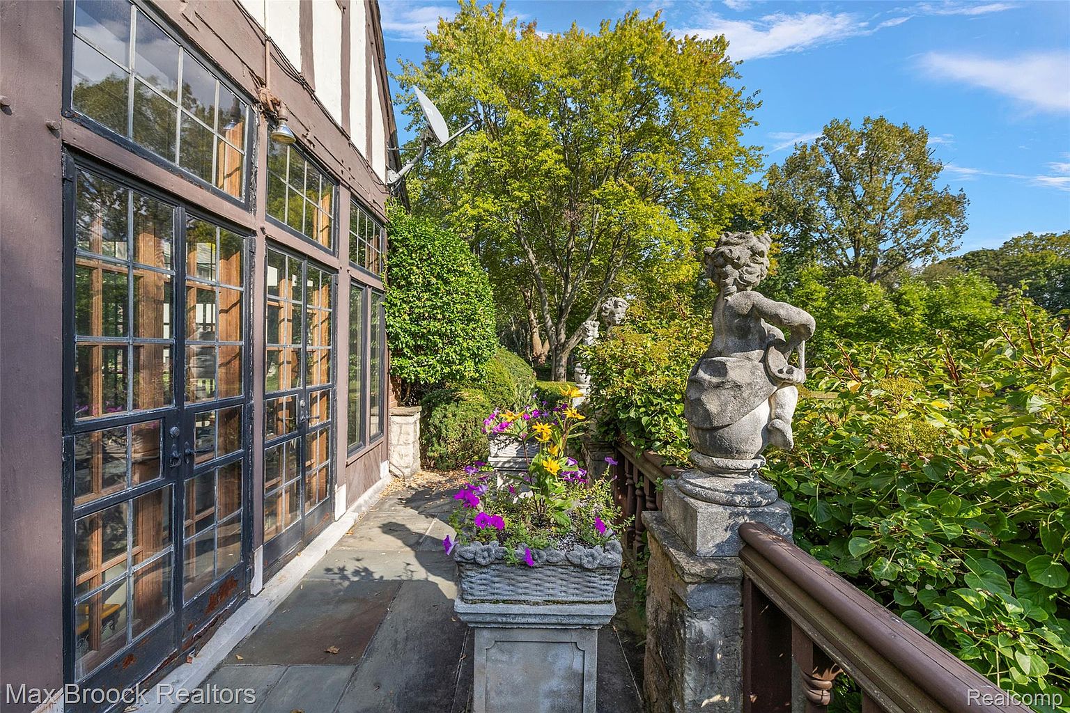 This image showcases a charming garden area adjacent to a building with large, multi-paned windows. A stone patio leads to a garden featuring a stone planter filled with colorful flowers and a decorative statue atop a stone pillar. Lush greenery and mature trees create a serene and inviting outdoor space.