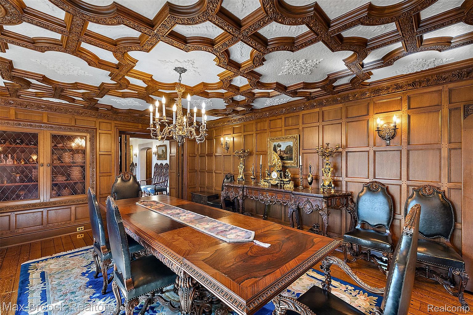 This is a luxurious dining room featuring ornate wood paneling, a coffered ceiling with intricate detailing, and a grand chandelier. A large, elaborately carved dining table is the centerpiece, surrounded by elegant chairs. The room exudes a sense of classic opulence and sophistication, perfect for formal gatherings and fine dining.