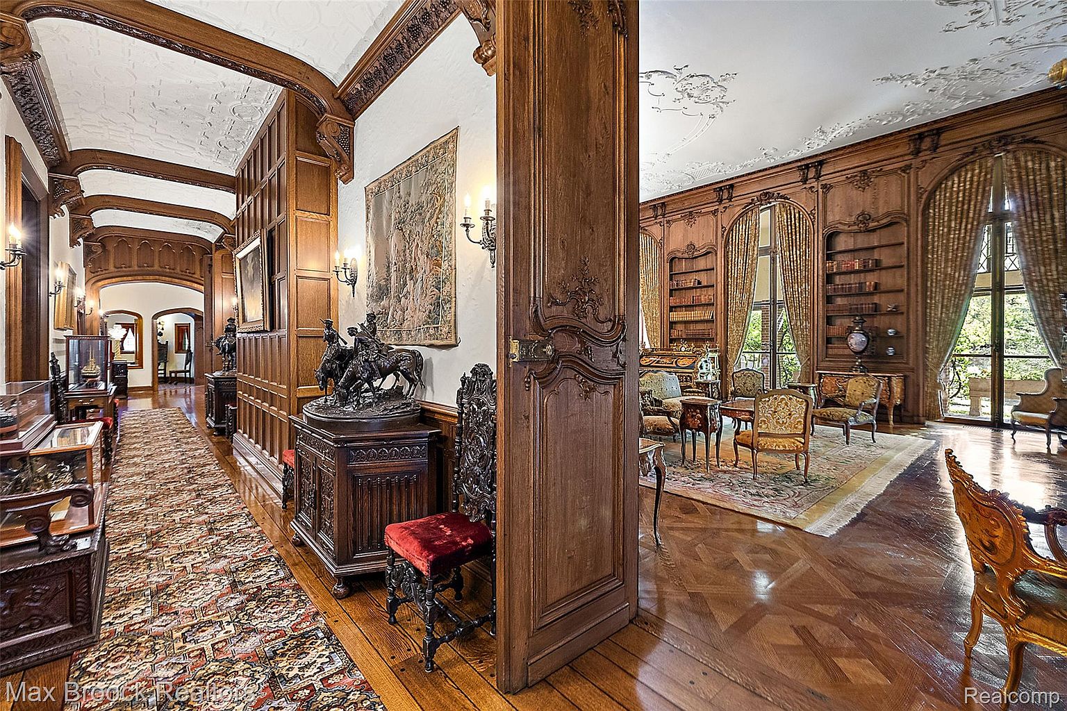 This image showcases a luxurious interior featuring a grand hallway leading into a richly decorated living room or library. The hallway is adorned with intricate wood paneling, a patterned rug, and antique furniture, while the adjacent room boasts floor-to-ceiling bookshelves, elegant seating, and large windows with ornate curtains. The overall impression is one of opulence and classic design, highlighting the home's architectural details and sophisticated ambiance.