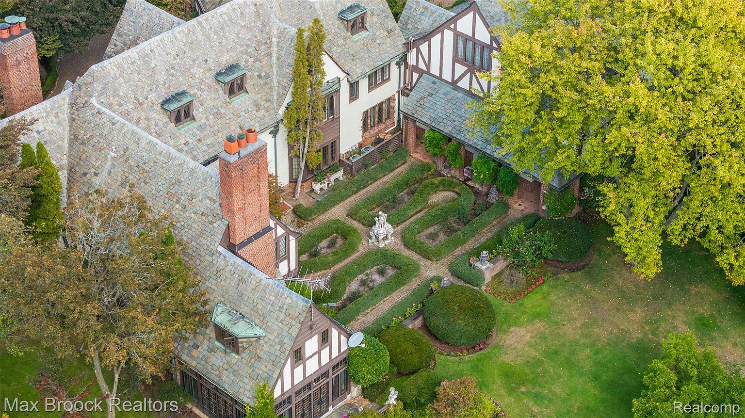 This aerial shot showcases an expansive estate with meticulously manicured gardens, featuring geometric hedges and a central fountain. The Tudor-style architecture is evident in the steeply pitched roofs, half-timbered details, and prominent brick chimneys. The property exudes a sense of timeless elegance and grandeur.