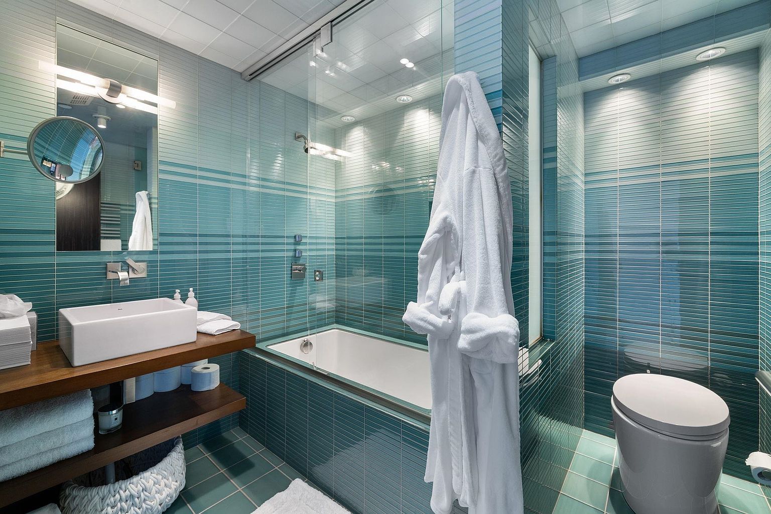 This is a primary bathroom featuring a modern design with blue-green tiled walls and floors. A white rectangular sink sits on a wooden shelf, complemented by a large mirror and modern lighting. A bathtub with a glass enclosure is visible, along with a white robe hanging nearby, and a toilet in a separate alcove. The overall impression is clean, contemporary, and spa-like.