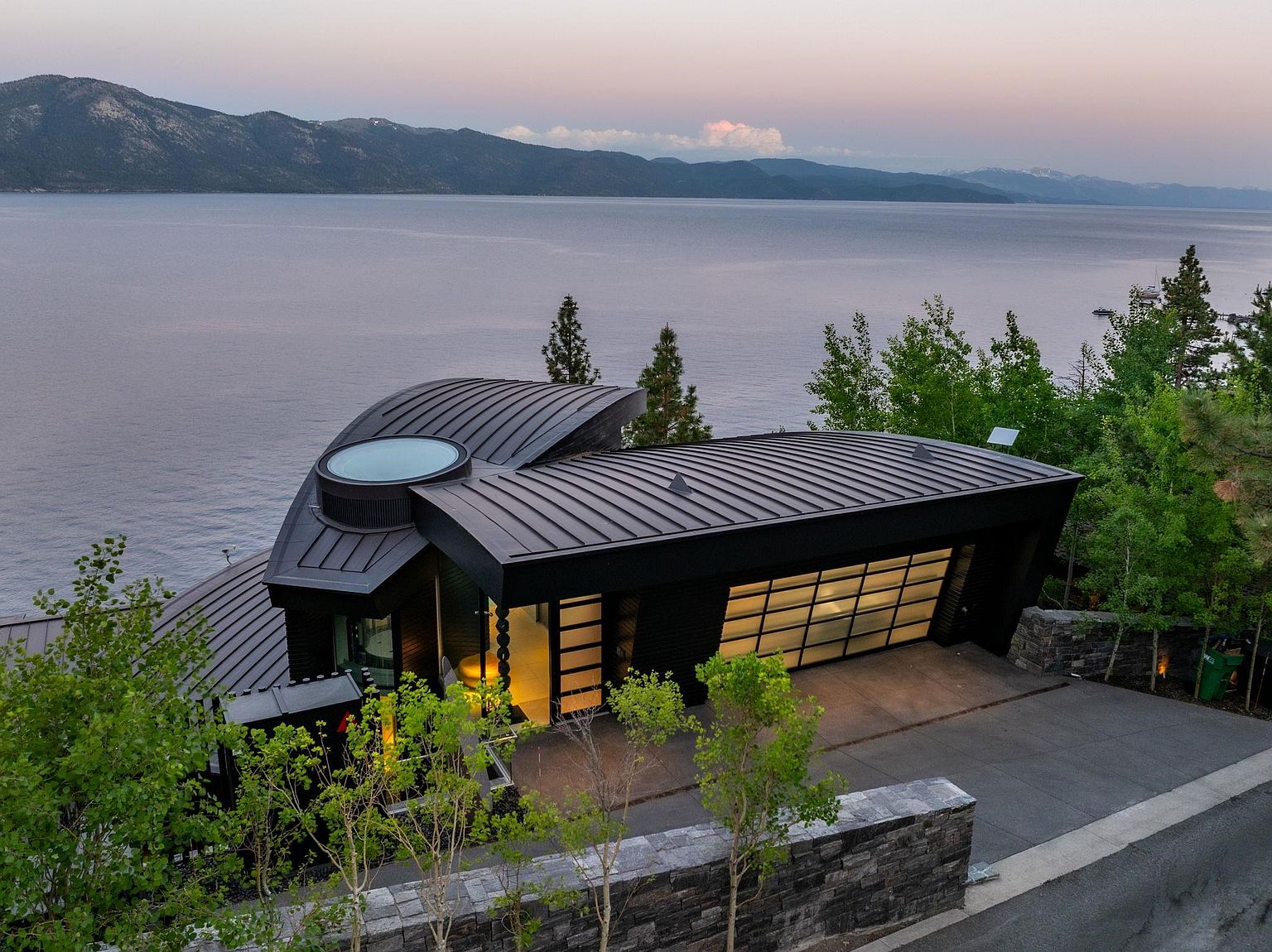 This aerial view showcases a modern architectural masterpiece nestled by a serene lake and mountains. The house features a unique curved roof, a glass-paneled garage door, and a stone retaining wall, blending seamlessly with the natural landscape. The overall impression is one of luxury and tranquility.