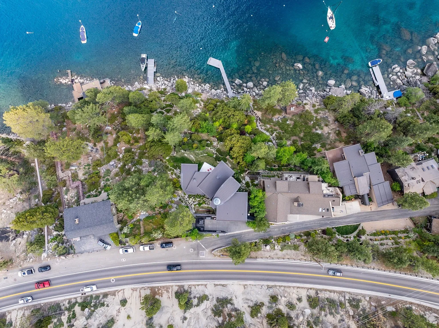 This aerial shot captures a collection of luxury homes nestled along a picturesque lakefront. The properties boast modern architectural designs with distinctive rooflines, complemented by lush greenery and private docks. The turquoise waters of the lake and the winding road adjacent to the properties enhance the exclusive and serene setting, appealing to discerning buyers seeking privacy and natural beauty.