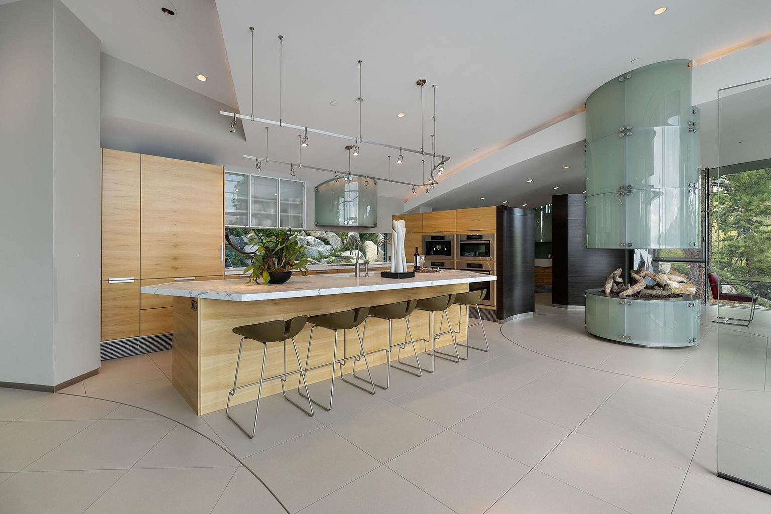 This is a spacious, modern kitchen featuring a large island with seating, light wood cabinetry, and a unique cylindrical fireplace. The kitchen is well-lit with a combination of recessed and pendant lighting, and the open layout creates a seamless flow into the adjacent living space. The design emphasizes clean lines and natural materials, creating a sophisticated and inviting atmosphere.