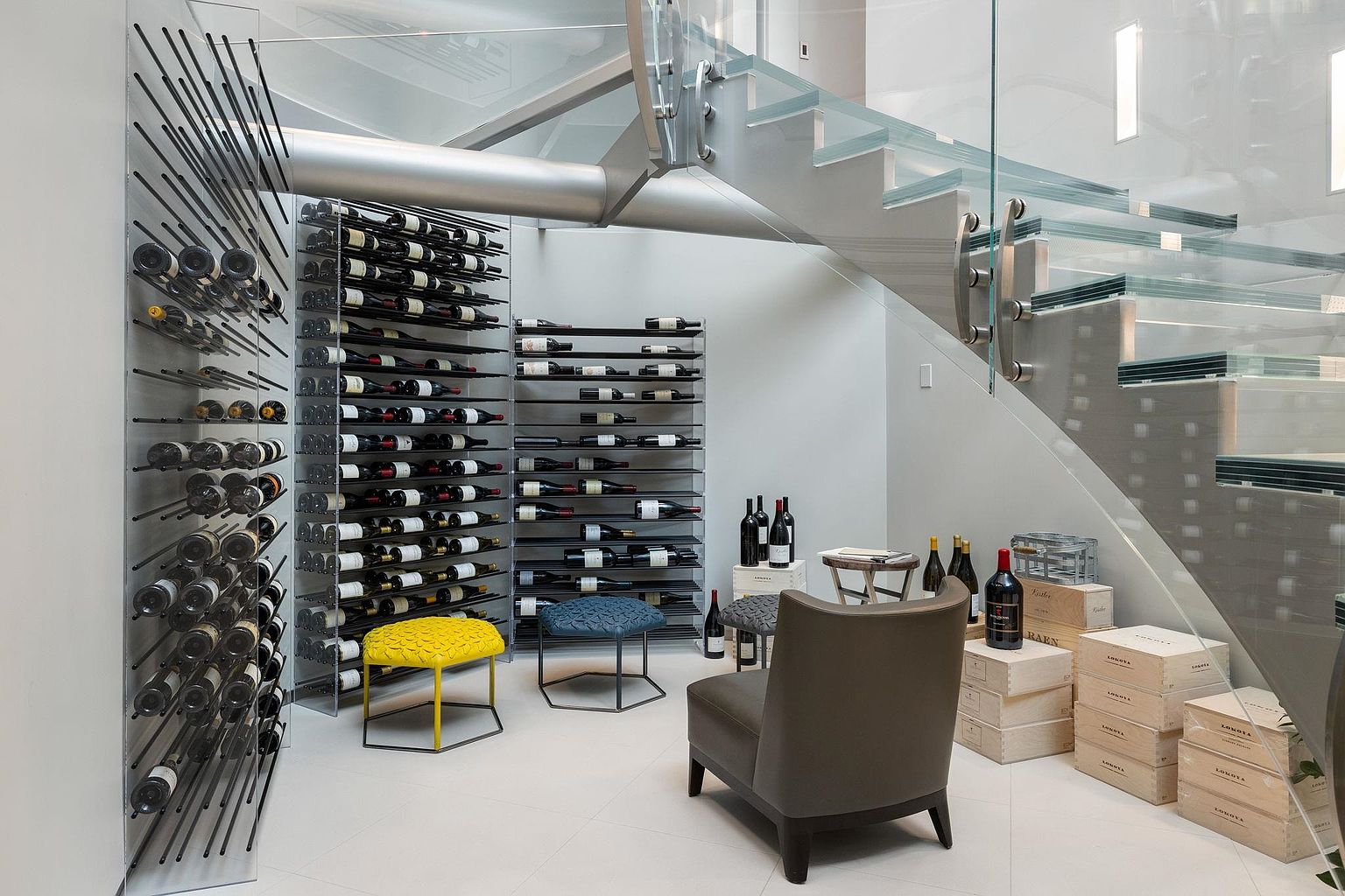This is an interior shot of a modern wine cellar, featuring sleek metal wine racks filled with bottles. A glass staircase adds a contemporary touch, while a comfortable chair and small stools provide seating. Wooden crates are stacked nearby, enhancing the cellar's aesthetic.