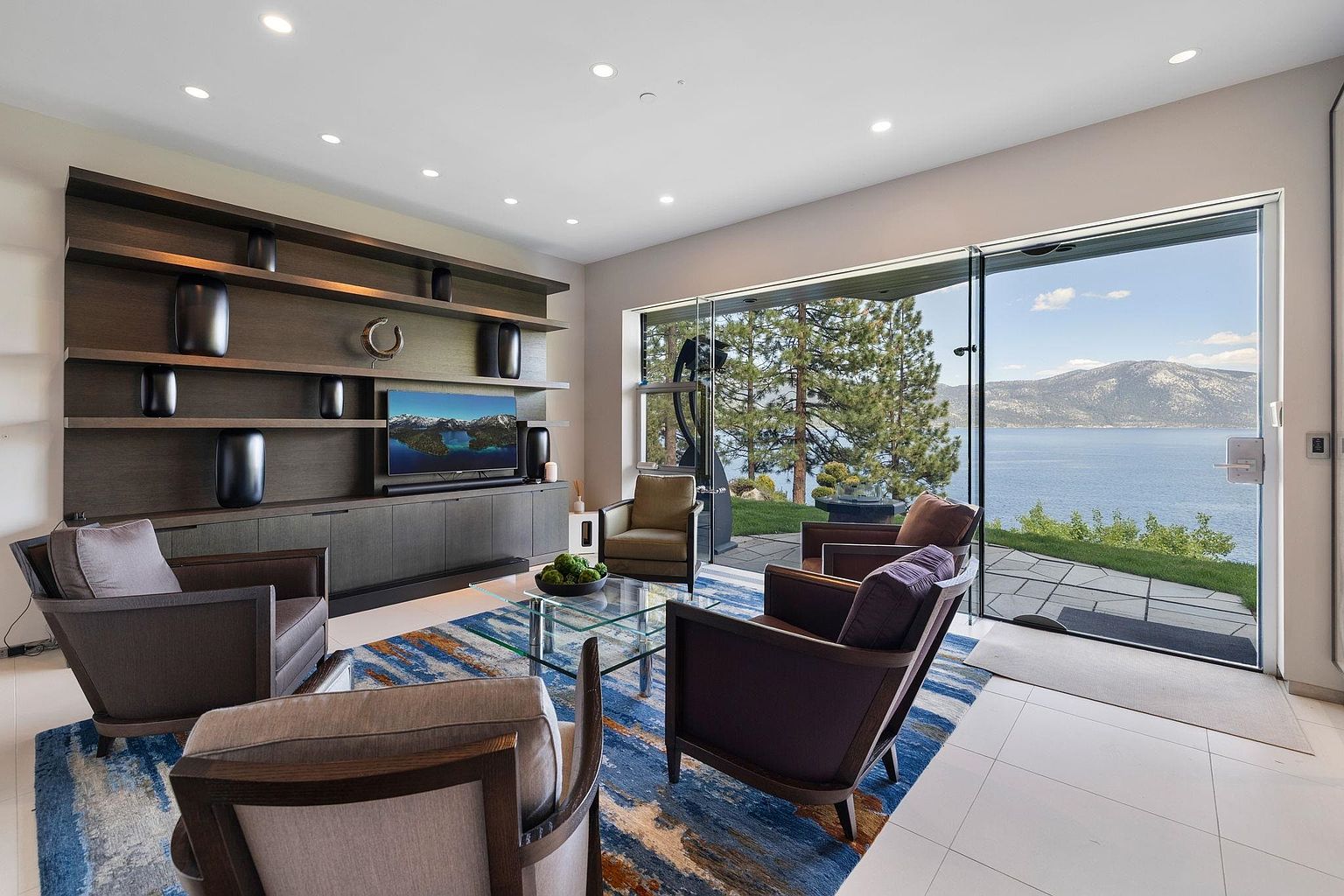 This is a modern living room with a stunning view of a lake and mountains through a large sliding glass door. The room features a built-in shelving unit with decorative items and a television, along with comfortable armchairs arranged around a glass coffee table on a blue and orange rug. The overall impression is one of luxury and relaxation, with a focus on the beautiful natural surroundings.