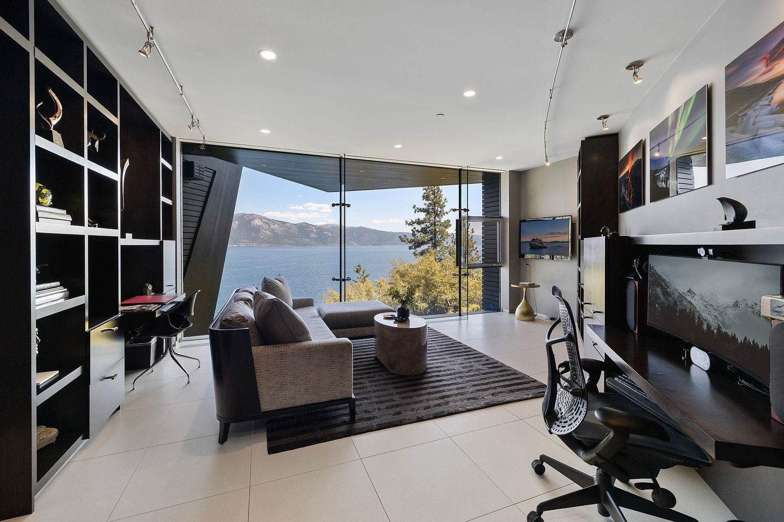 This is a modern office/study with a stunning lake view. The room features a built-in bookshelf, a sleek desk area with a large monitor, and a comfortable seating area with a sectional sofa and a round coffee table. The large windows provide ample natural light and showcase the beautiful scenery, creating a serene and productive workspace.