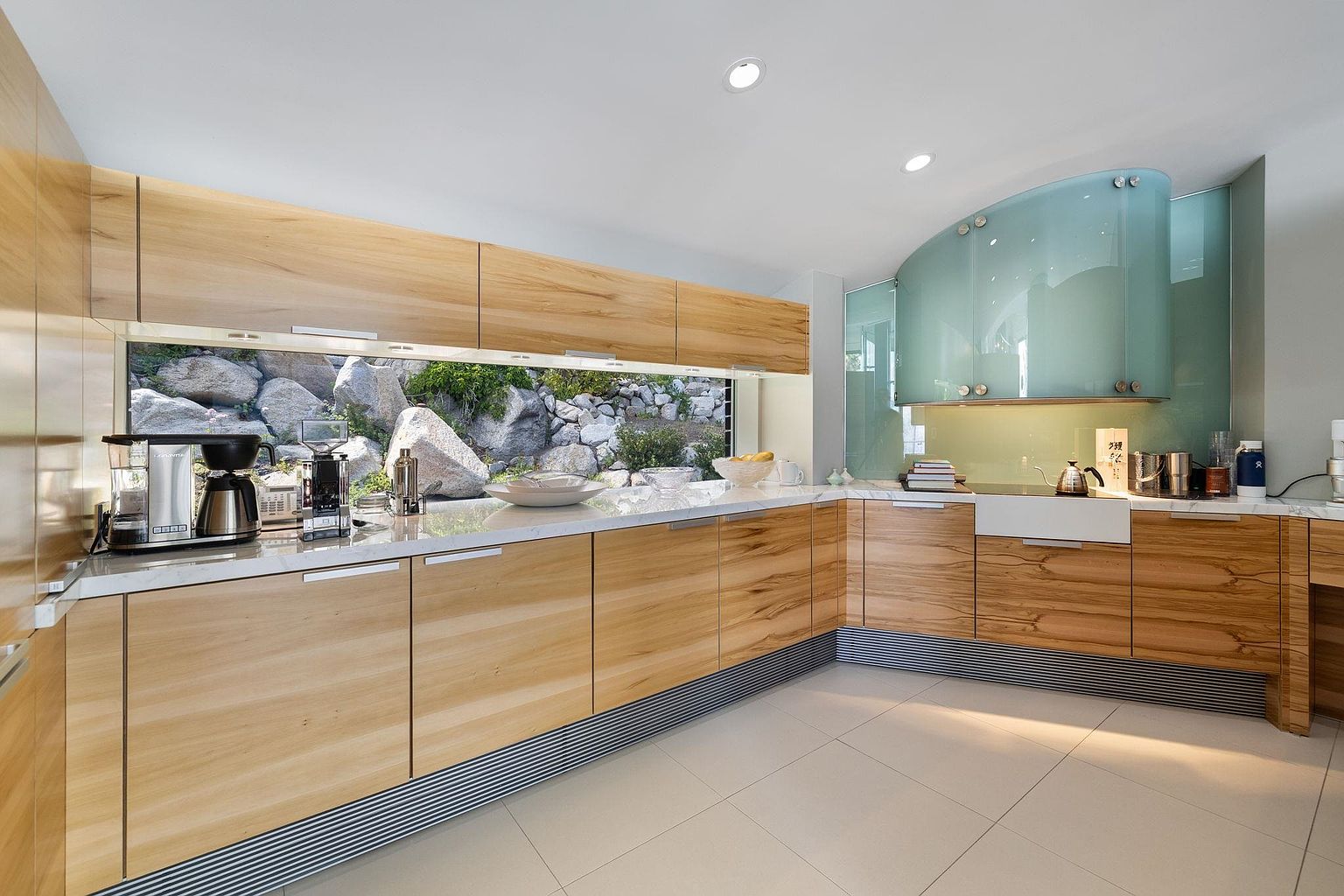 This is a modern kitchen featuring light wood cabinetry with sleek, minimalist hardware and a marble countertop. A unique window provides a view of a rock garden, bringing natural light and an organic element into the space. The kitchen also features a curved, frosted glass backsplash and white tile flooring, creating a bright and airy atmosphere.