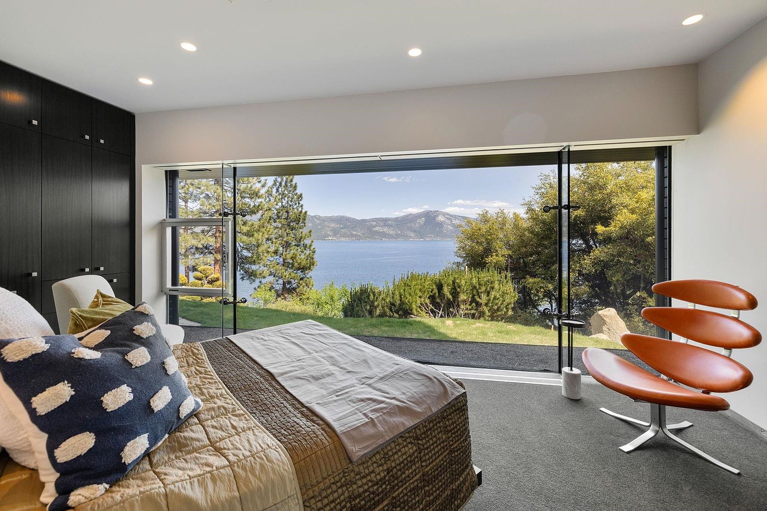 This primary bedroom boasts a stunning view of a lake and mountains through a large picture window, creating a serene and luxurious atmosphere. The room features a modern design with a dark wardrobe, a unique leather chair, and a comfortable bed with stylish pillows and bedding. The gray carpet adds a touch of elegance and comfort to the space.