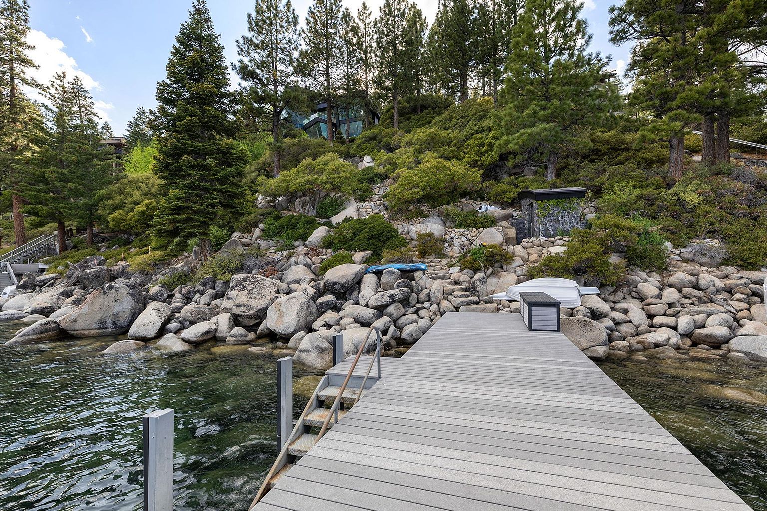 The image showcases a waterfront property featuring a wooden dock extending into clear water. The dock has metal stairs leading down to the water, and the shoreline is lined with large rocks and lush greenery. In the background, a modern house is nestled among the trees on the hillside, creating a serene and luxurious atmosphere.