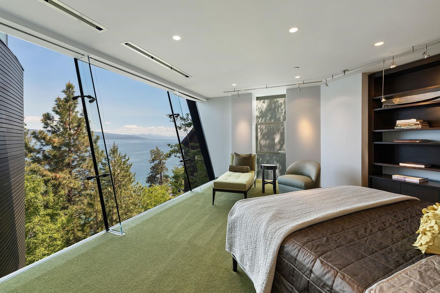 This primary bedroom boasts a stunning panoramic view through floor-to-ceiling windows, showcasing a serene lake and lush greenery. The room features a chaise lounge, a comfortable armchair, and a bed with a quilted coverlet, creating a relaxing and luxurious atmosphere. A built-in bookshelf adds a touch of sophistication and functionality to the space.