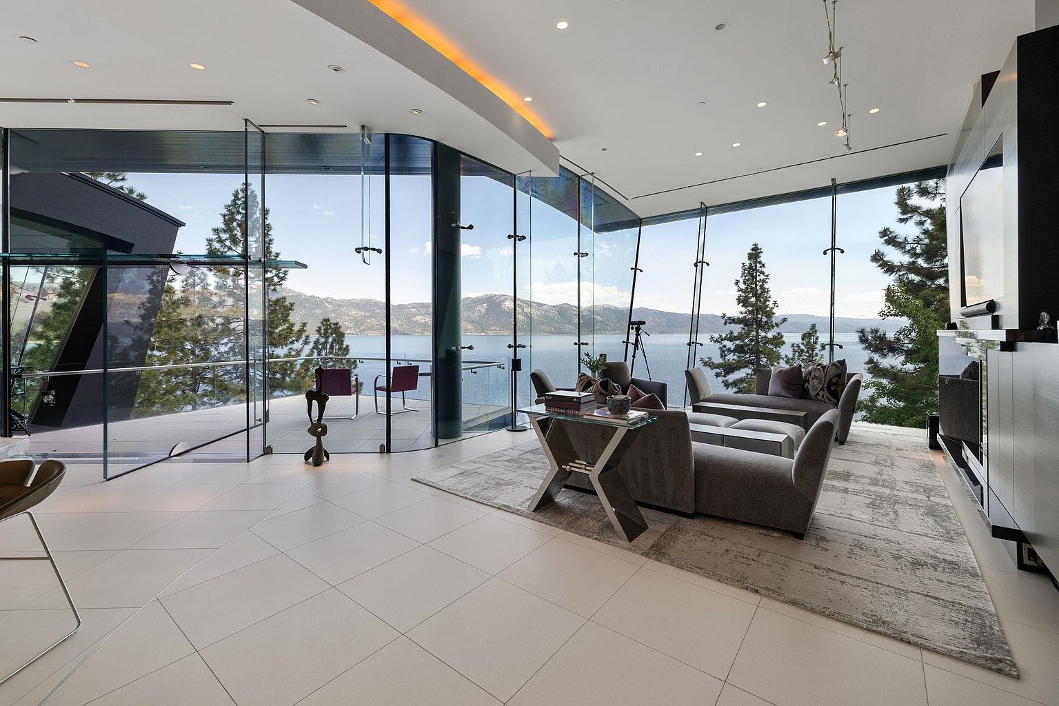 This is a luxurious living room with floor-to-ceiling windows offering panoramic views of a lake and mountains. The room features modern furniture, including a gray sofa and armchairs, arranged on a patterned rug over white tile flooring. A sleek entertainment center and contemporary lighting fixtures add to the room's sophisticated ambiance.