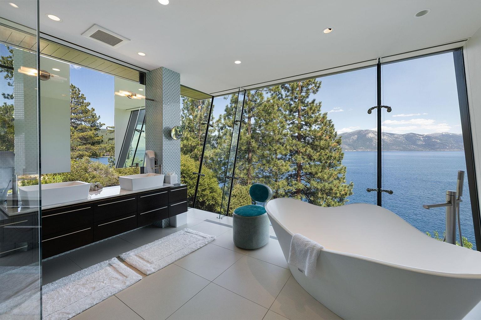 This is a luxurious primary bathroom with floor-to-ceiling windows offering stunning views of a lake and mountains. The bathroom features a modern freestanding bathtub, a double vanity with sleek dark wood cabinets, and a separate glass-enclosed shower. The design emphasizes natural light and a seamless connection to the outdoor scenery, creating a spa-like atmosphere.
