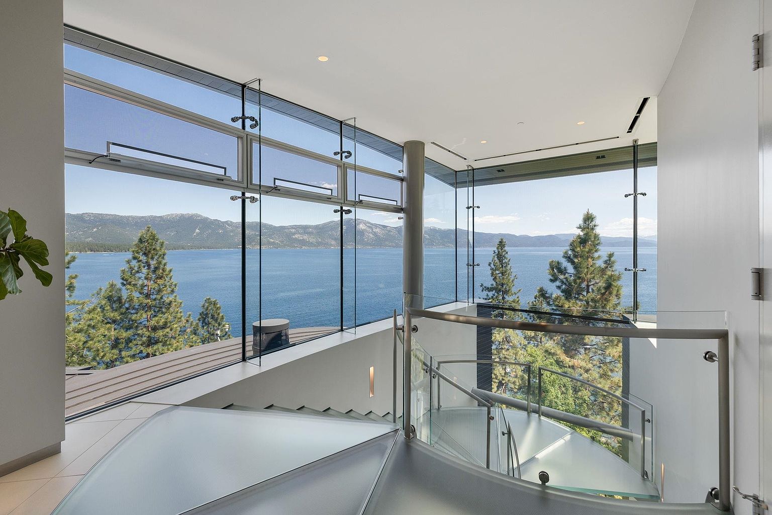 This interior shot showcases a modern staircase with glass railings and frosted treads, offering a stunning view of a lake and mountains through floor-to-ceiling windows. The design emphasizes natural light and open space, creating a luxurious and airy atmosphere. The staircase appears to be a spiral staircase.