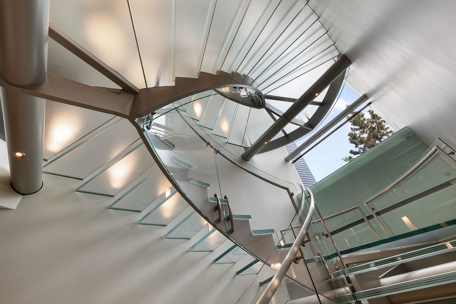 This image showcases a modern, architectural staircase with glass steps and railings, creating a light and airy feel. The design emphasizes clean lines and a minimalist aesthetic, with metal supports adding a touch of industrial chic. The view is from below, looking up the spiral staircase, emphasizing its height and design.