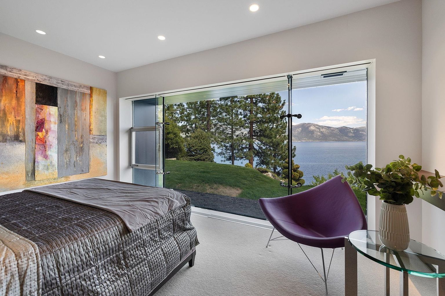 This primary bedroom features a large window with a view of a lake and mountains, bringing the outdoors in. The room is decorated with modern art and a stylish purple chair, complemented by a glass side table with a plant. The bed is neatly made with a textured gray comforter, creating a serene and inviting atmosphere.