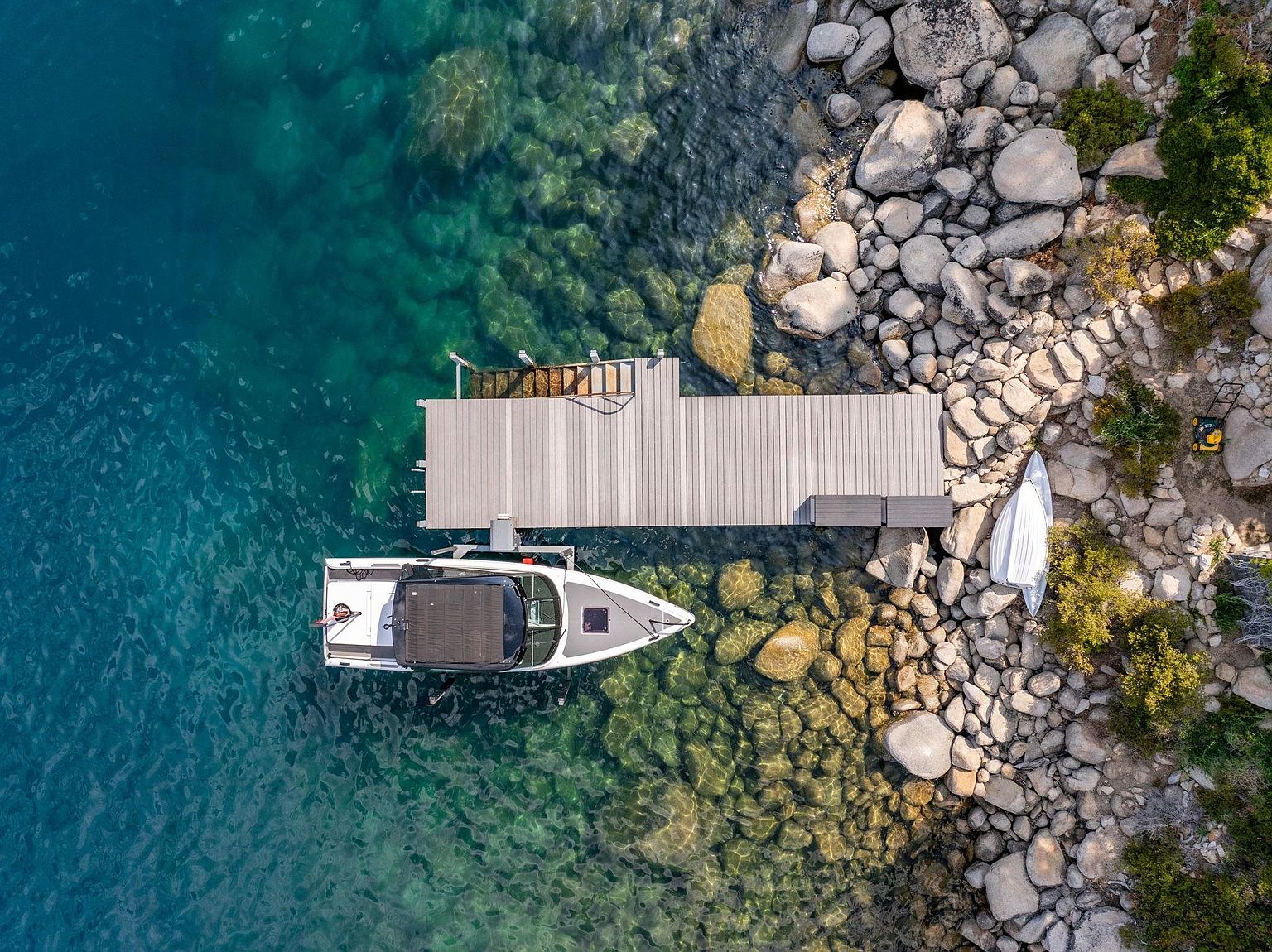 This aerial shot showcases a pristine waterfront property with crystal-clear turquoise water. A modern boat is docked at a wooden pier, adjacent to a rocky shoreline with lush greenery. The scene evokes a sense of luxury and tranquility, highlighting the property's prime location and access to recreational water activities.