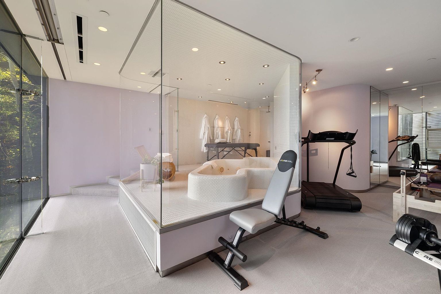 This interior shot showcases a luxurious home gym and spa area. The space features a modern design with a glass-enclosed spa area including a jacuzzi and massage table, complemented by exercise equipment such as a treadmill and weight bench. The color palette is soft and calming, creating a serene and upscale atmosphere.