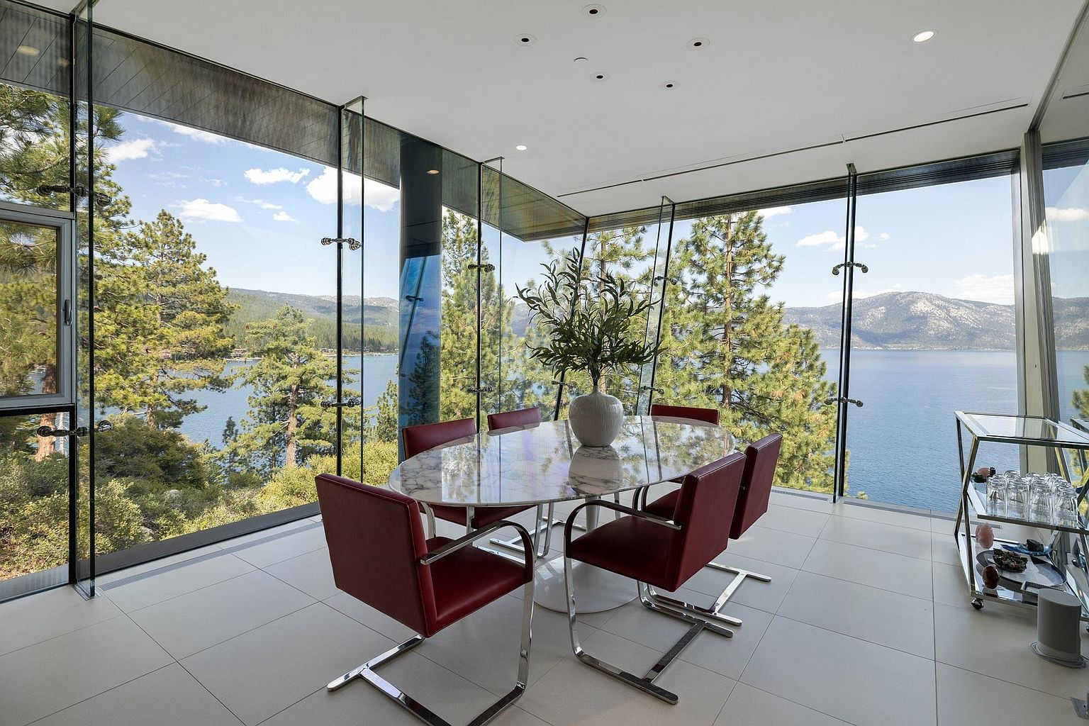 This is a bright and modern dining room with floor-to-ceiling windows offering stunning views of a lake and surrounding trees. The room features a marble-topped oval dining table surrounded by six red leather chairs with chrome frames. A sleek bar cart is visible in the corner, adding a touch of sophistication.