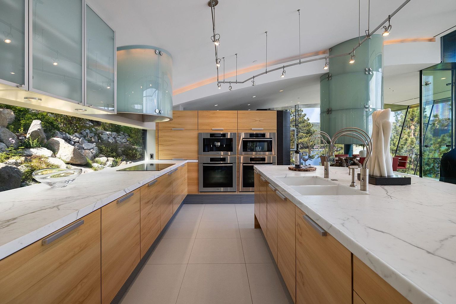This is a modern kitchen with light wood cabinetry and marble countertops. The kitchen features high-end stainless steel appliances and a large window with a view of the outdoors. The design incorporates unique architectural elements, such as a cylindrical glass structure, creating a bright and open space.