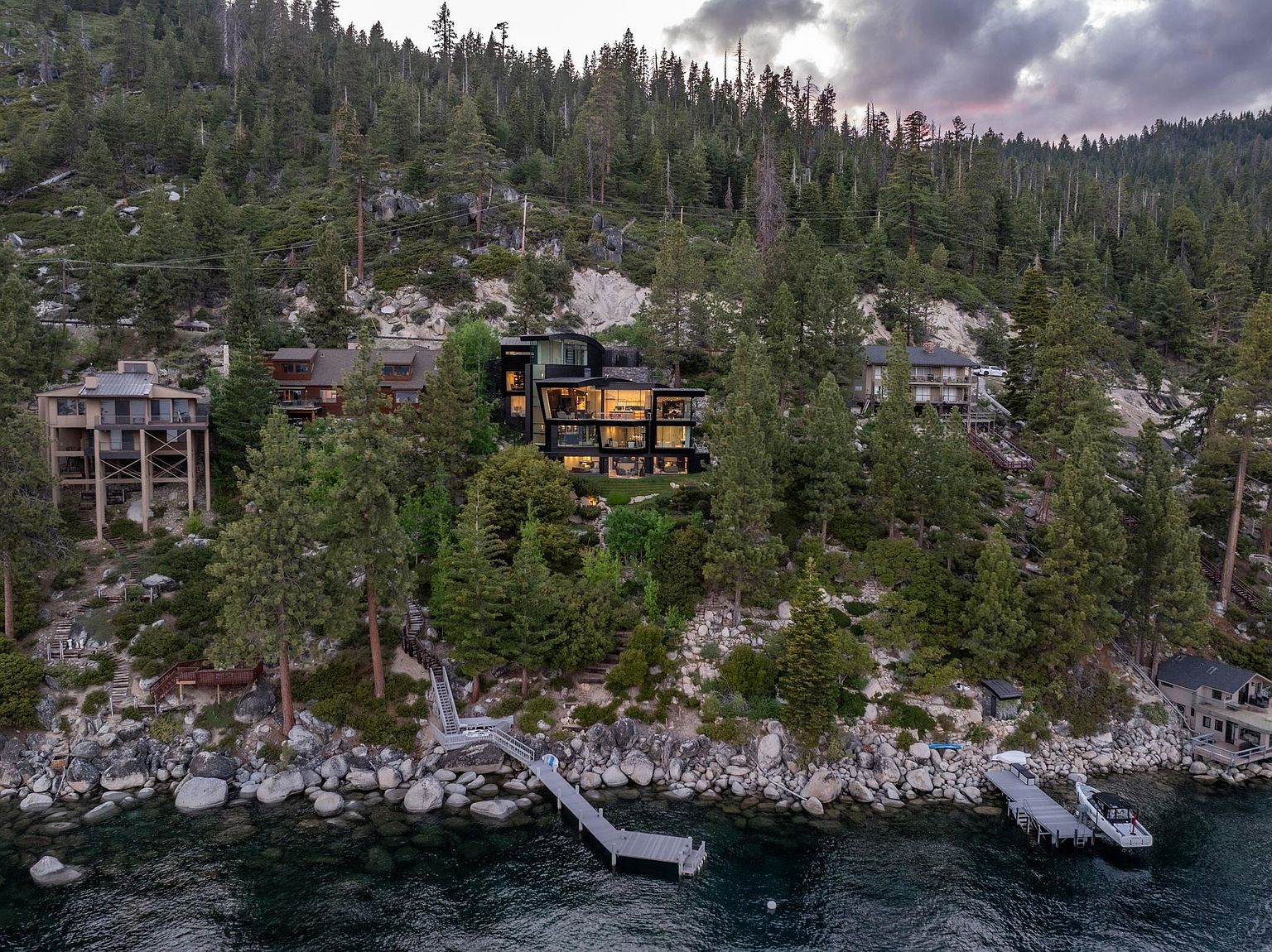This aerial view showcases a stunning multi-story modern home nestled on a wooded hillside overlooking a lake. The property features a private dock and boat lift, emphasizing its waterfront access and recreational opportunities. The surrounding landscape is lush with mature trees, creating a sense of privacy and natural beauty.
