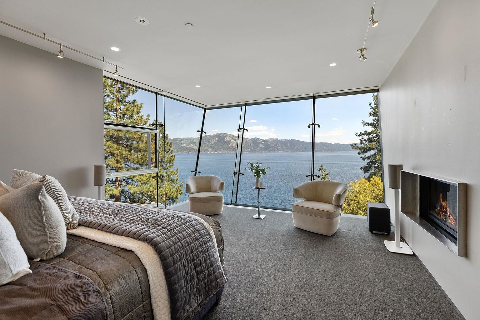 This is a luxurious primary bedroom with a stunning view of a lake and mountains through floor-to-ceiling windows. The room features a modern fireplace, two comfortable armchairs, and a plush bed with high-end bedding. The overall aesthetic is contemporary and serene, emphasizing the breathtaking natural surroundings.