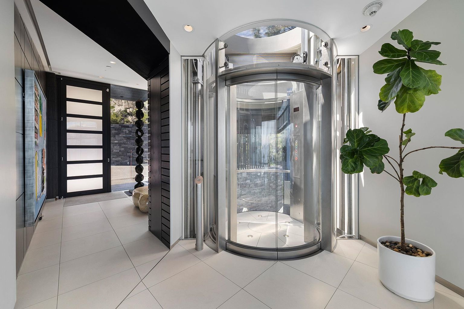 This interior shot showcases a modern hallway featuring a unique glass elevator as a focal point. The hallway is brightly lit with white tile flooring, and a large potted plant adds a touch of nature. The perspective is from a medium distance, highlighting the architectural details and the seamless integration of the elevator into the home's design.