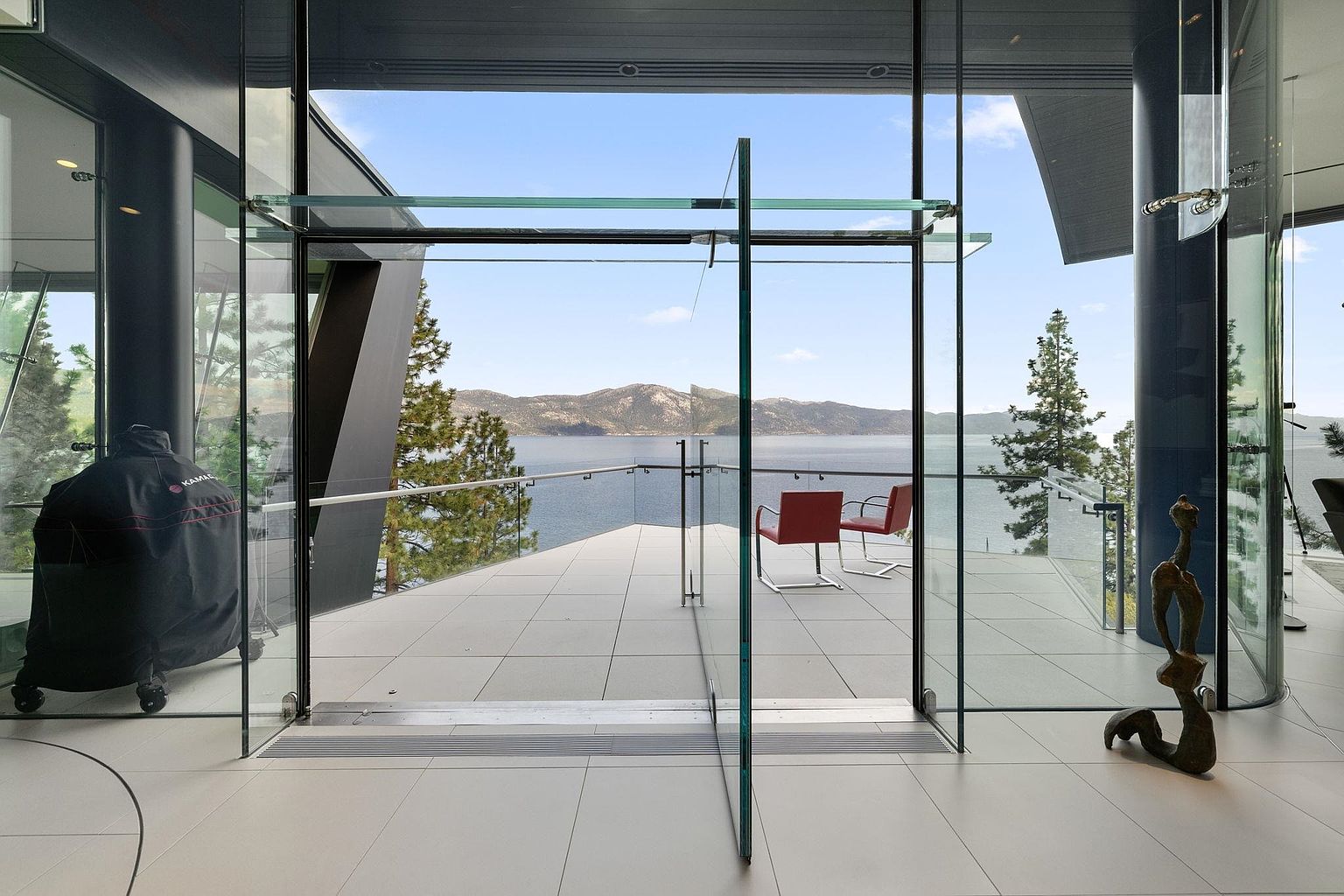 This image showcases a modern patio or balcony with a stunning lake view. The space features sleek, light-colored tiling and is furnished with two red chairs. Large glass doors and panels create a seamless transition between the interior and exterior, emphasizing the open and airy design of the property.