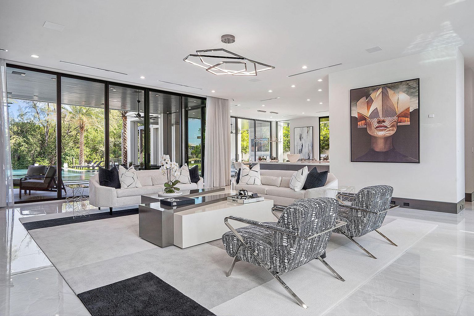 This is a luxurious living room featuring two white sofas with decorative pillows, two patterned armchairs, and a modern coffee table. Large windows offer views of the outdoor pool and greenery, while a striking piece of art adorns the wall. The room is well-lit with a unique geometric chandelier and recessed lighting, creating a bright and inviting atmosphere.