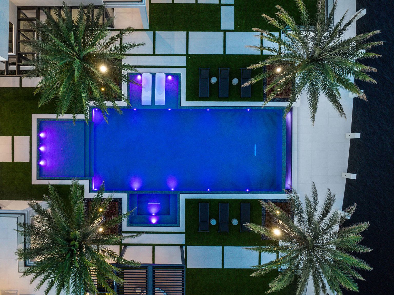 This aerial view showcases a luxurious backyard featuring a rectangular pool illuminated with blue lighting, surrounded by meticulously maintained green spaces and modern white tile pathways. Palm trees add a tropical touch, while lounge chairs and small tables offer relaxation spots. The overall design exudes sophistication and tranquility, perfect for upscale outdoor living.