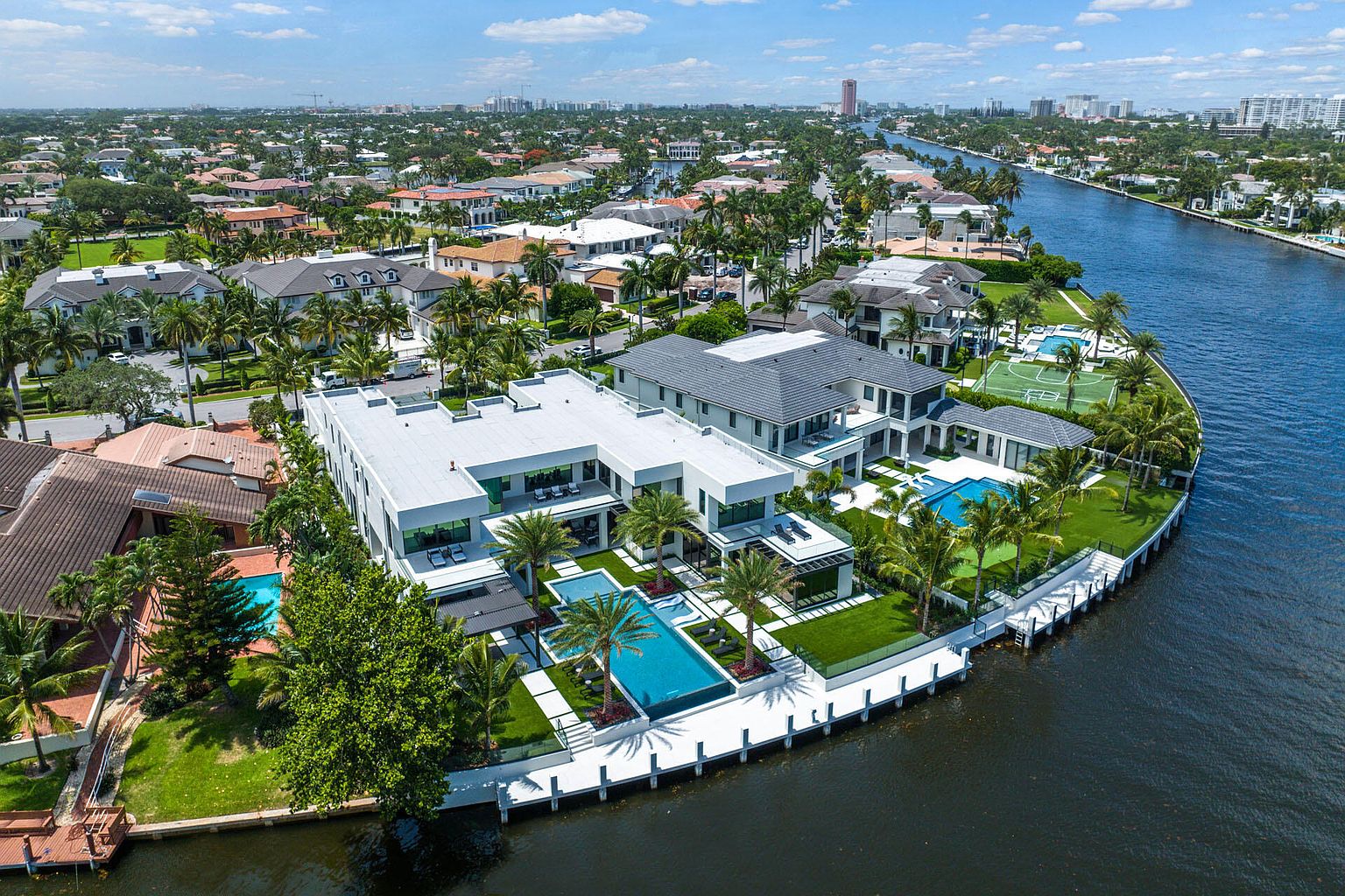 This aerial shot showcases a luxurious waterfront property featuring a modern architectural design with clean lines and a flat roof. The house boasts multiple pools, lush landscaping with palm trees, and a private dock along the waterway. The surrounding neighborhood consists of similar high-end homes, creating an exclusive and desirable residential area.