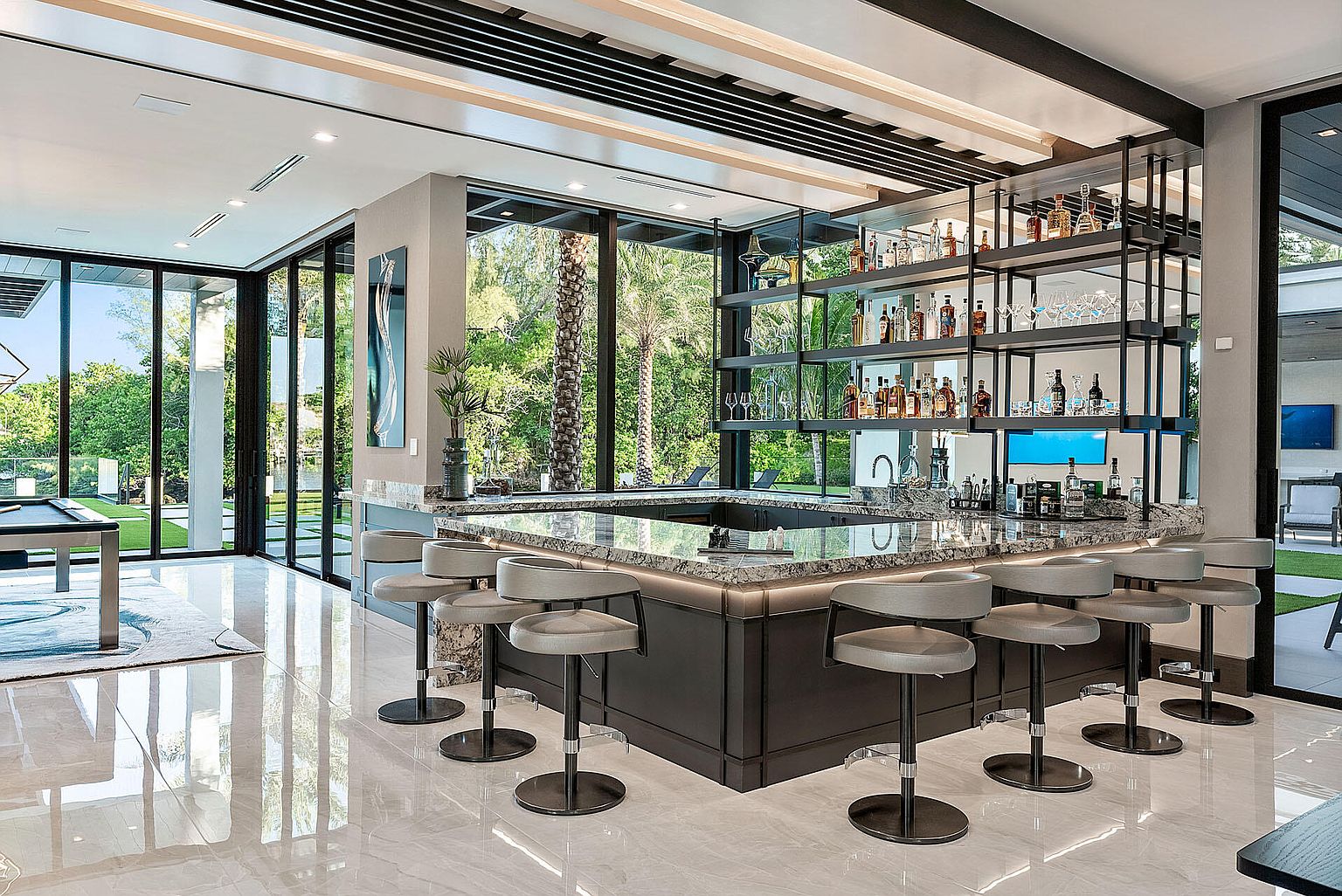 This interior shot showcases a modern living room featuring a stylish bar area. The bar boasts a granite countertop, dark cabinetry, and open shelving displaying various bottles and glassware. Large windows provide ample natural light and views of the exterior, while contemporary bar stools offer seating.