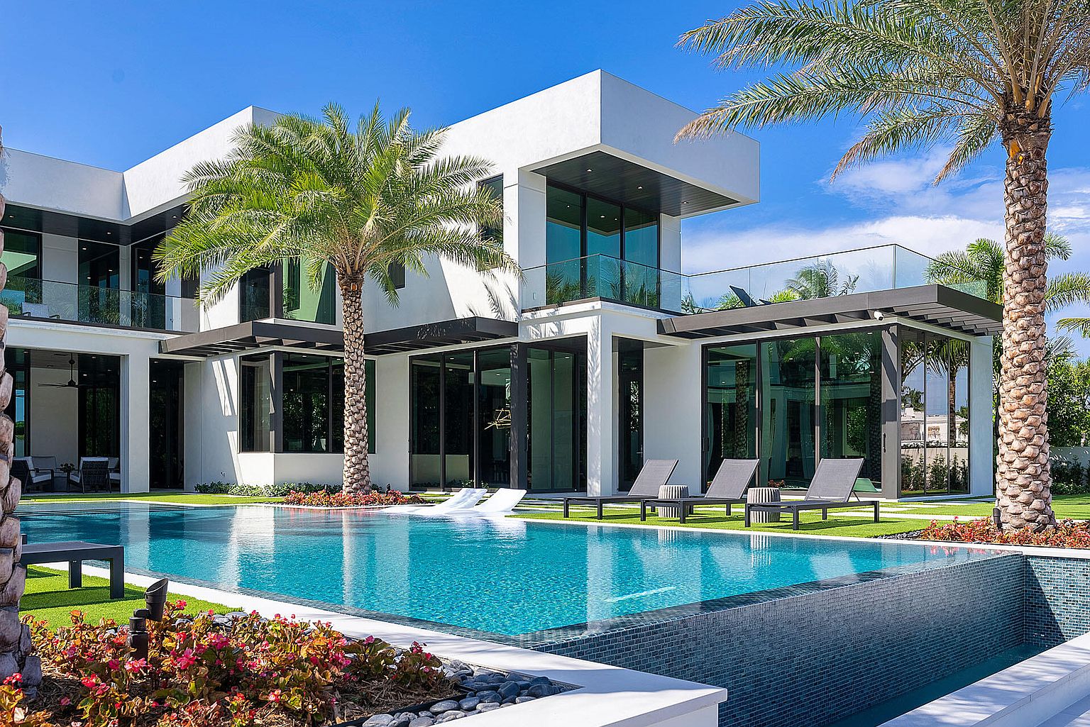 This image showcases a luxurious modern home with a stunning backyard pool area. The pool features a sleek, contemporary design with clear blue water, surrounded by well-manicured green grass, palm trees, and stylish lounge chairs. The architectural style of the house is modern with clean lines and large windows, creating a seamless indoor-outdoor living experience.