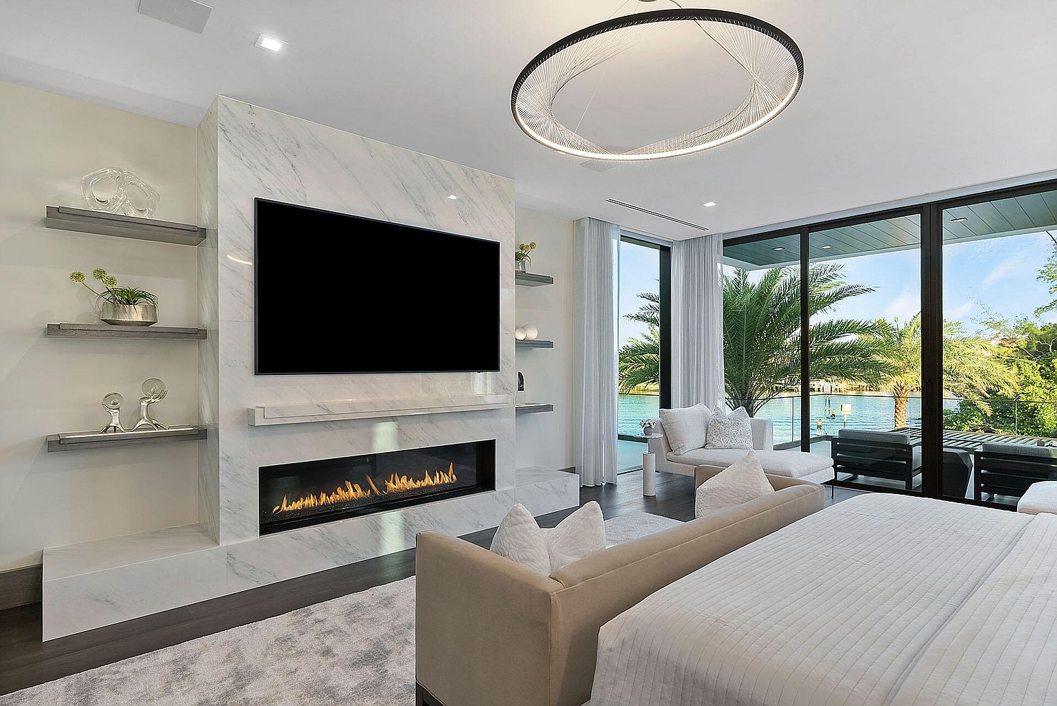 This is a luxurious primary bedroom featuring a modern fireplace with a large screen television above it. The room has floor-to-ceiling windows offering a view of the water and palm trees, and a large circular light fixture hangs from the ceiling. The overall impression is one of high-end comfort and contemporary design.