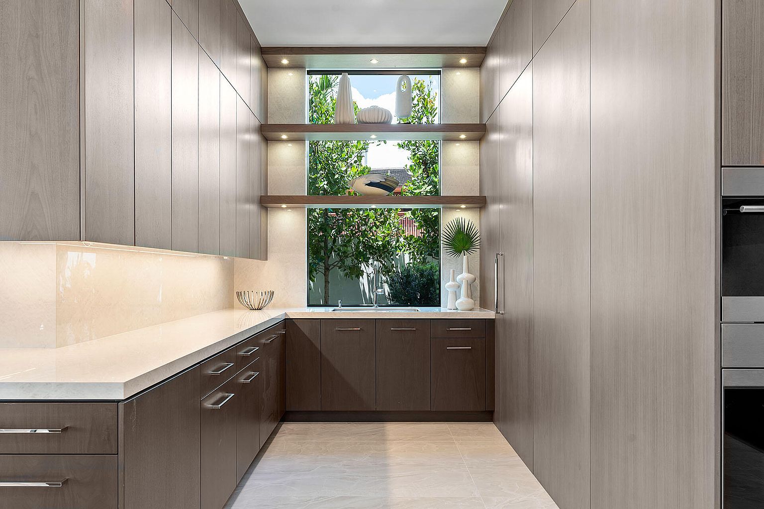 This is a modern kitchen featuring sleek, handleless cabinetry in two tones: a light wood grain for the tall cabinets and a darker wood for the lower cabinets. A unique window design with built-in shelving offers natural light and a view of greenery, while under-cabinet lighting illuminates the countertop. The overall impression is clean, contemporary, and well-organized.