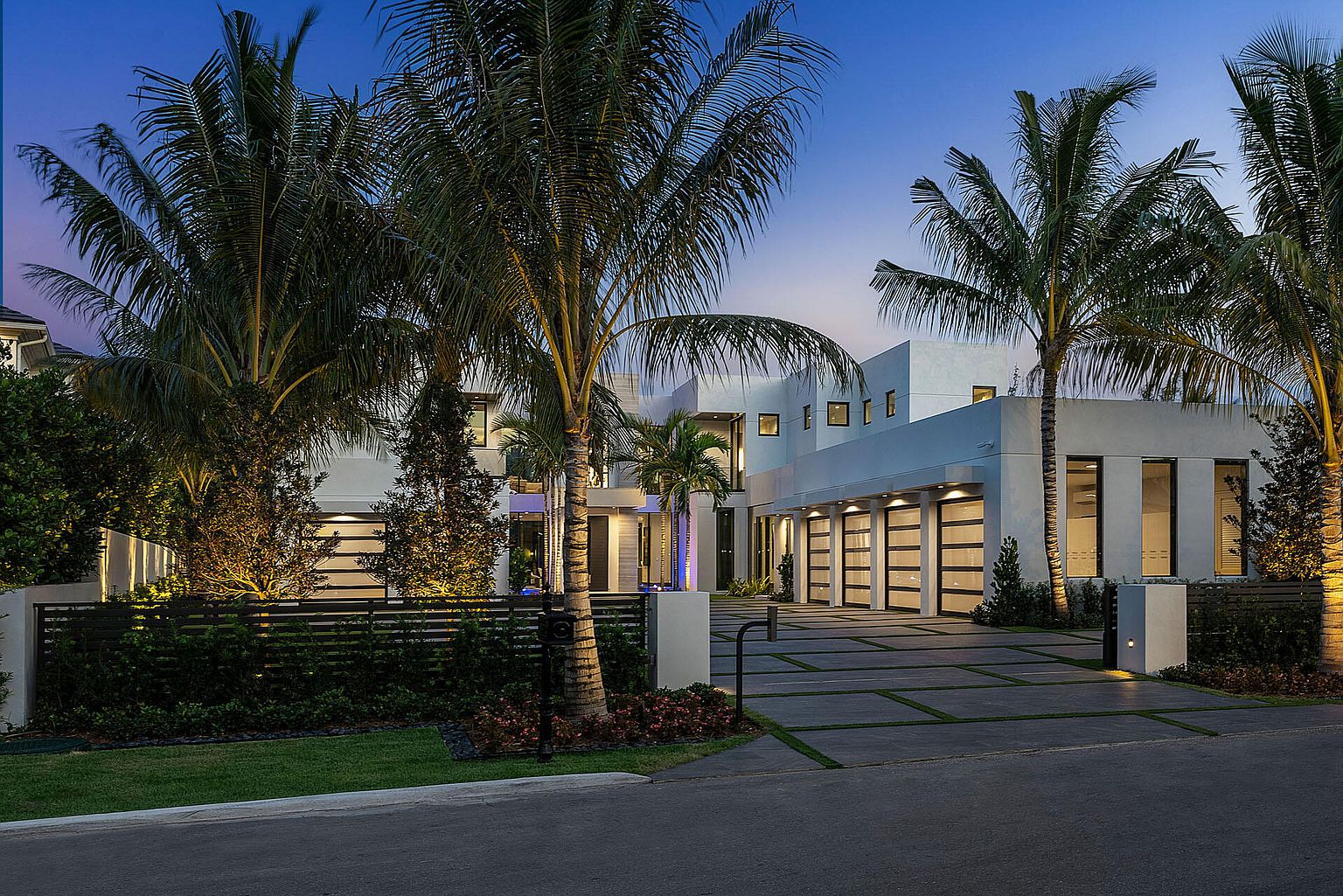 This is a front exterior view of a modern, luxurious home. The house features a white facade, multiple garage doors with black frames, and several palm trees in the front yard. The driveway is paved with concrete and grass accents, and the overall impression is one of upscale, contemporary living.