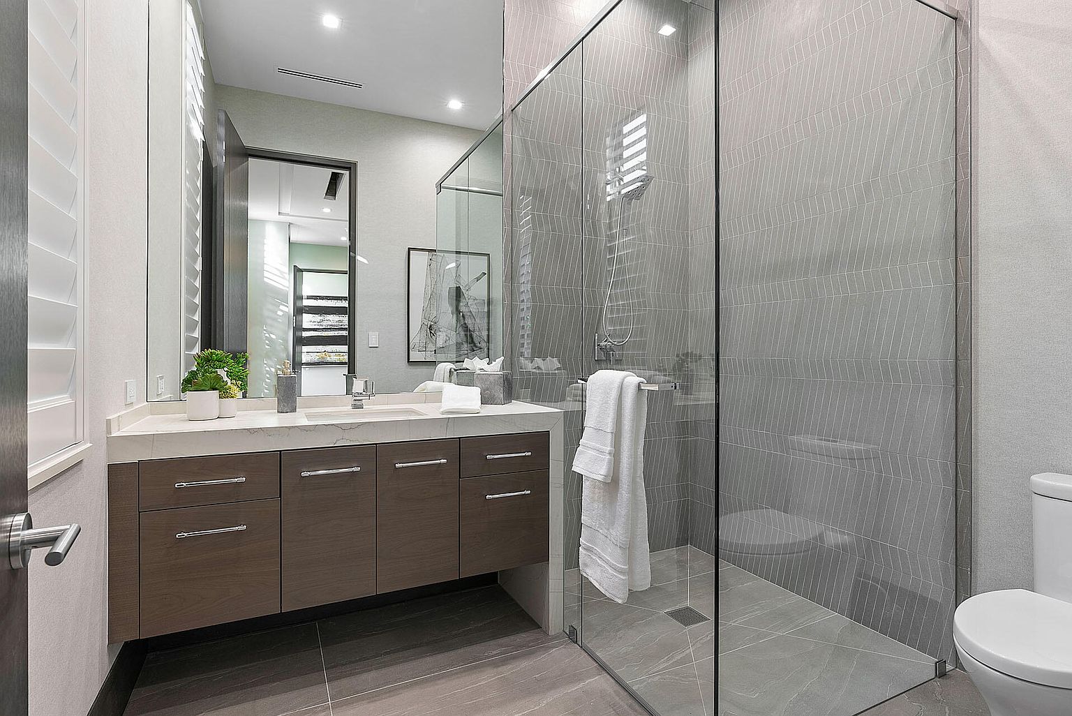 This is a modern bathroom featuring a floating vanity with dark wood cabinets and a white countertop. A large mirror hangs above the sink, reflecting the light and adding depth to the space. The shower is enclosed with glass panels and features gray patterned tiles, while a toilet sits adjacent to the shower area. The overall impression is clean, contemporary, and well-designed.