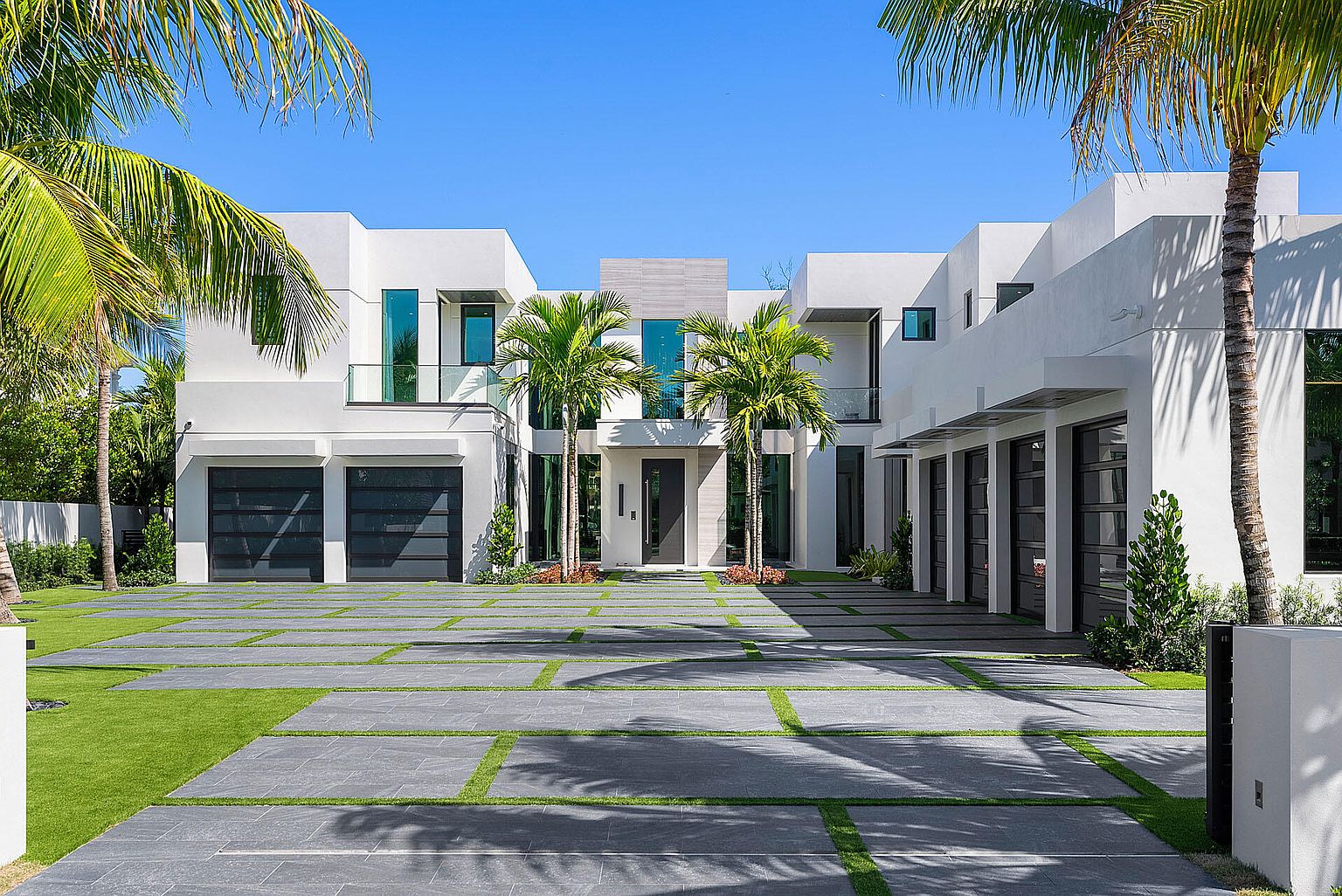 This is a front exterior view of a modern, two-story home with a flat roof and a symmetrical design. The house is painted white and features dark-framed windows and garage doors. The driveway is paved with large gray stones separated by strips of green grass, and palm trees are planted in the front yard, adding a tropical touch to the property.