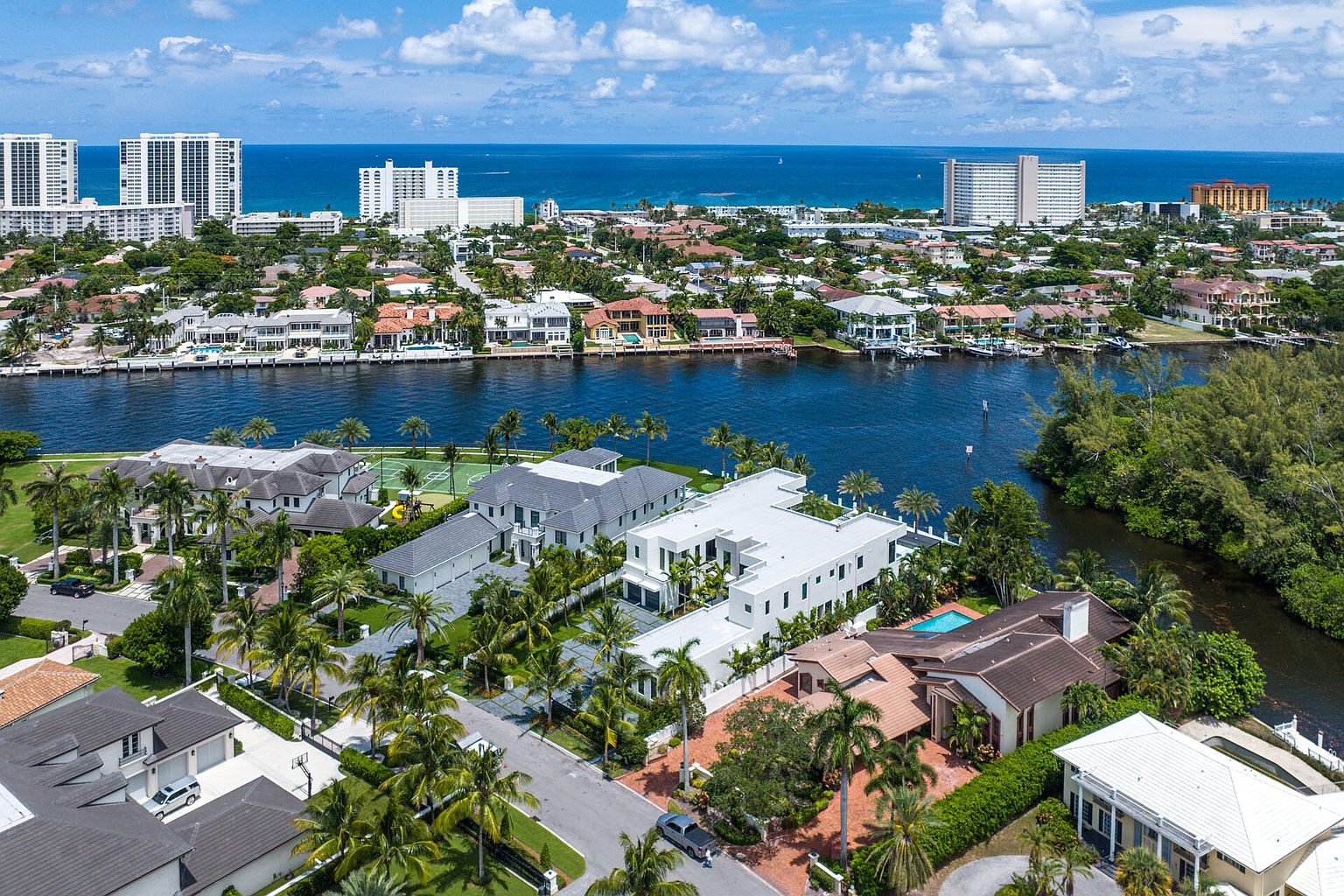 This aerial shot showcases a luxurious waterfront property with a modern architectural design, surrounded by lush landscaping and palm trees. The property features a large pool, a tennis court, and direct access to the waterway, offering stunning views of the ocean and the surrounding upscale neighborhood. The overall impression is one of opulence and exclusivity.