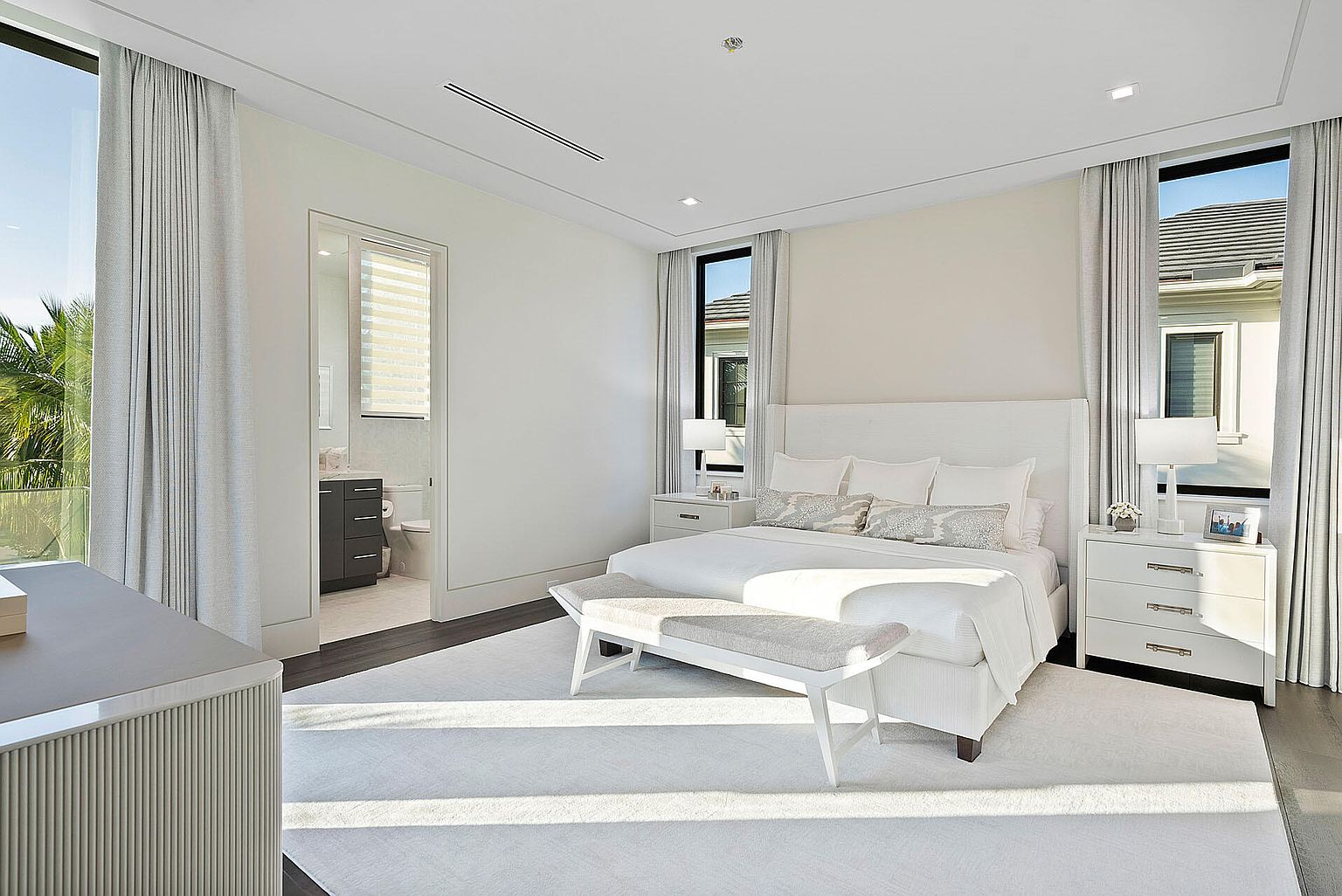 This is a bright and airy primary bedroom featuring a large bed with white linens and decorative pillows, flanked by matching nightstands and lamps. The room has a clean, modern aesthetic with neutral tones, hardwood floors, and large windows with sheer curtains that let in plenty of natural light. A bench sits at the foot of the bed, and a doorway leads to an ensuite bathroom.