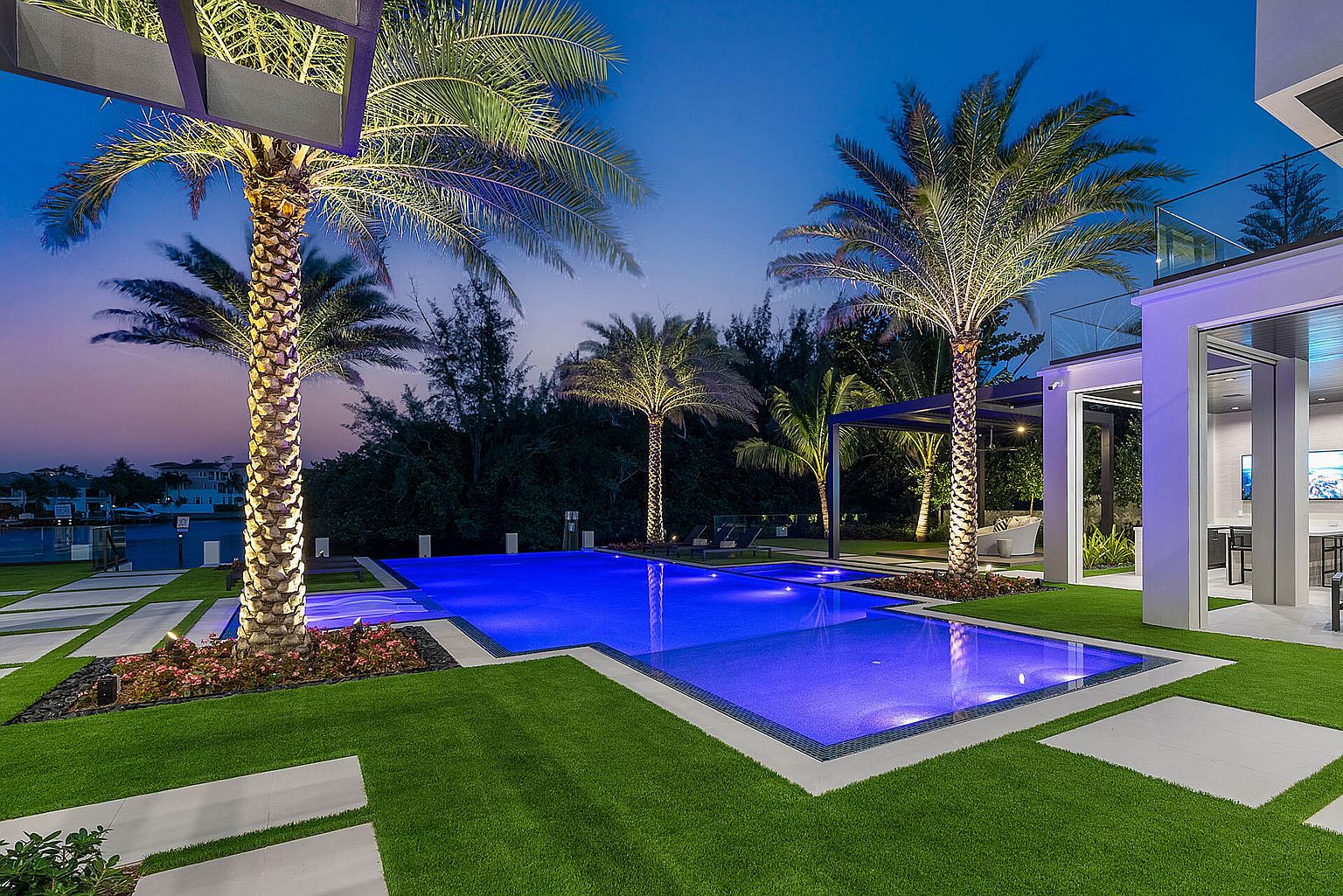 This image showcases a luxurious backyard pool area at dusk. The rectangular pool is illuminated with blue lighting, surrounded by manicured green grass and palm trees. The modern architecture of the house is visible in the background, creating an inviting and upscale outdoor living space.