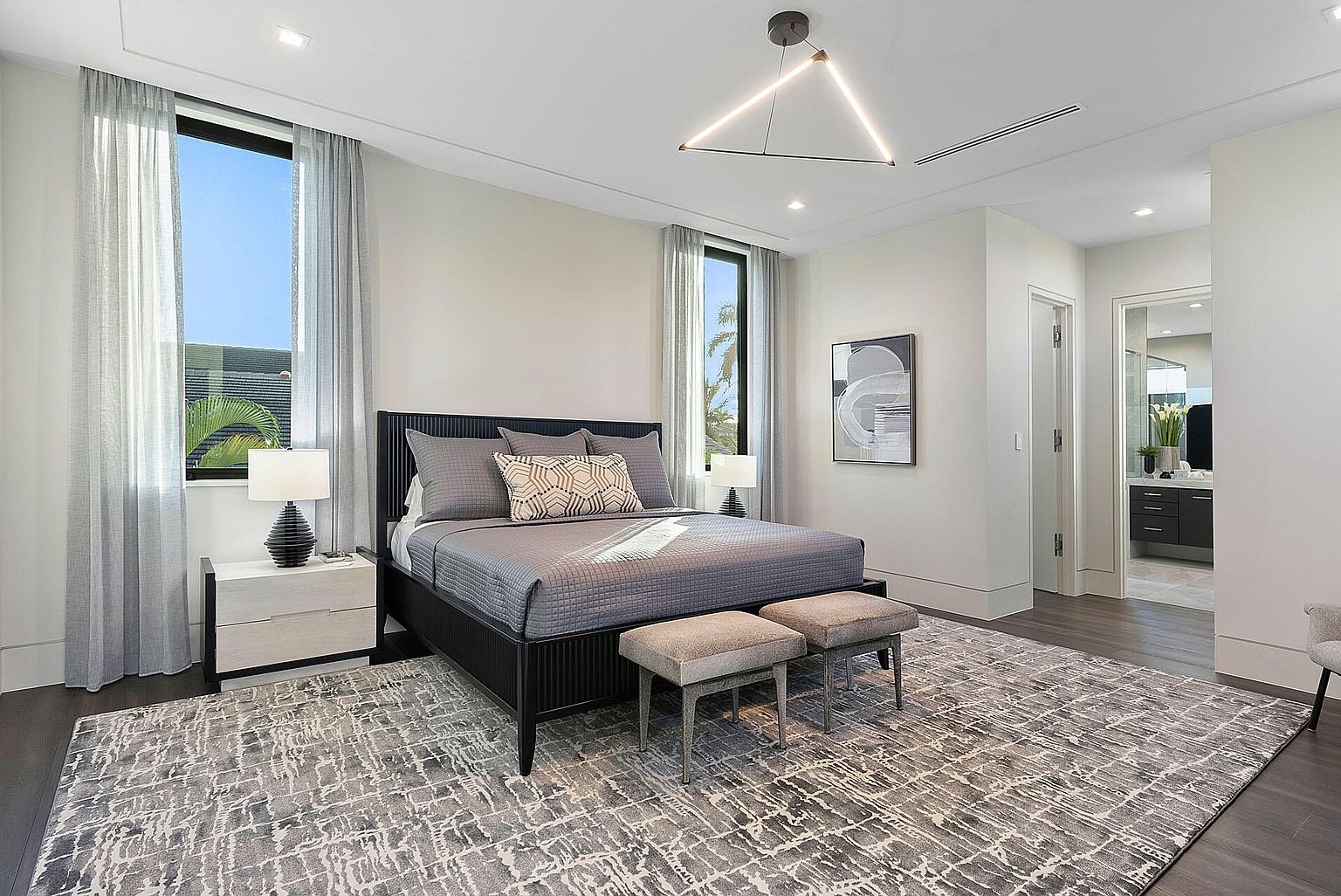This is a primary bedroom featuring a large bed with a gray quilted coverlet and decorative pillows, flanked by matching nightstands and lamps. The room has large windows with sheer curtains, allowing natural light to fill the space, and a modern geometric light fixture hangs from the ceiling. The overall aesthetic is contemporary and serene, with a neutral color palette and a spacious feel.
