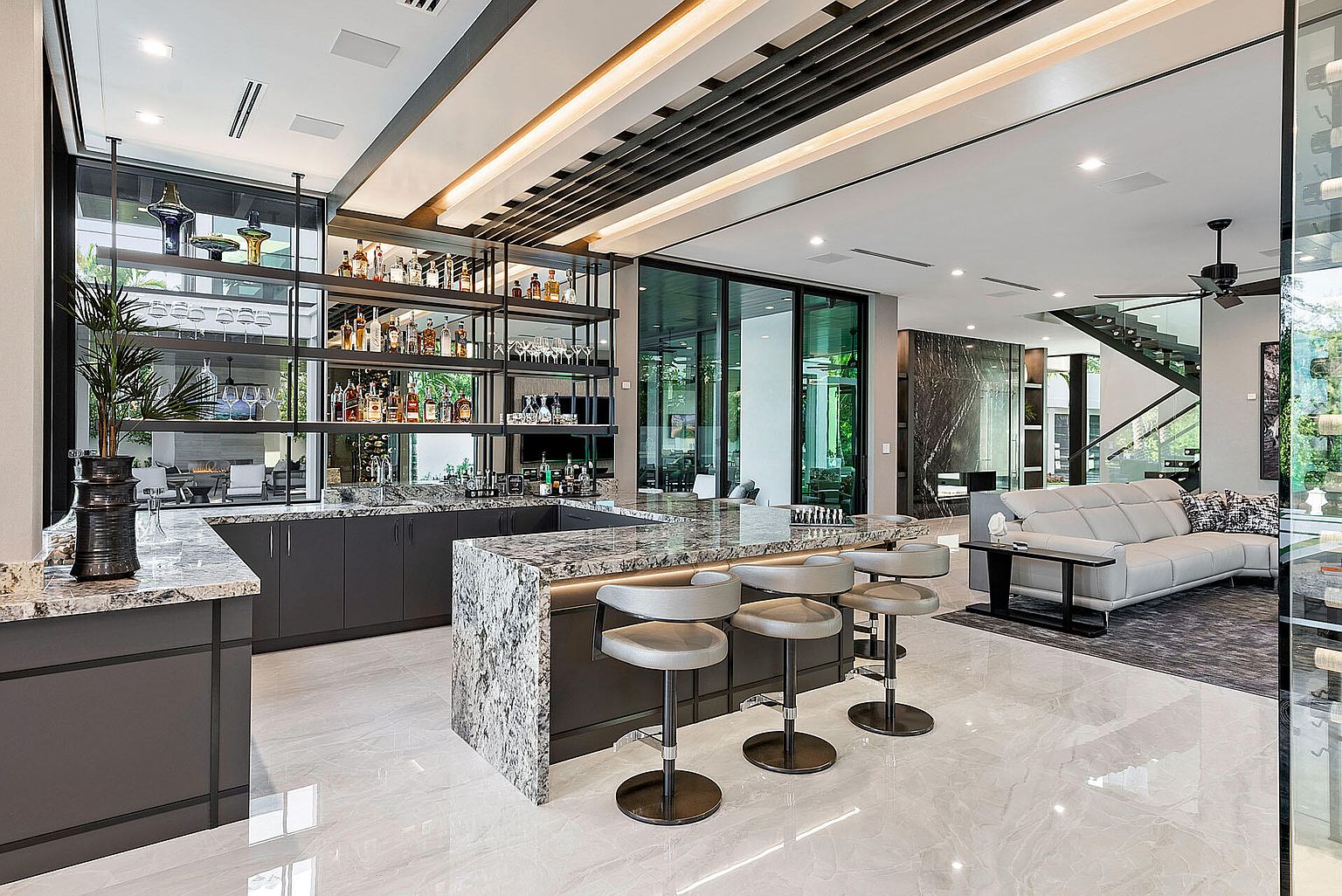 This interior shot showcases a modern living space with a stylish bar area featuring open shelving, granite countertops, and seating. The living room includes a sleek gray sofa, a dark rug, and a staircase in the background, all contributing to a luxurious and contemporary ambiance. Large sliding glass doors provide ample natural light and a view to the outside.