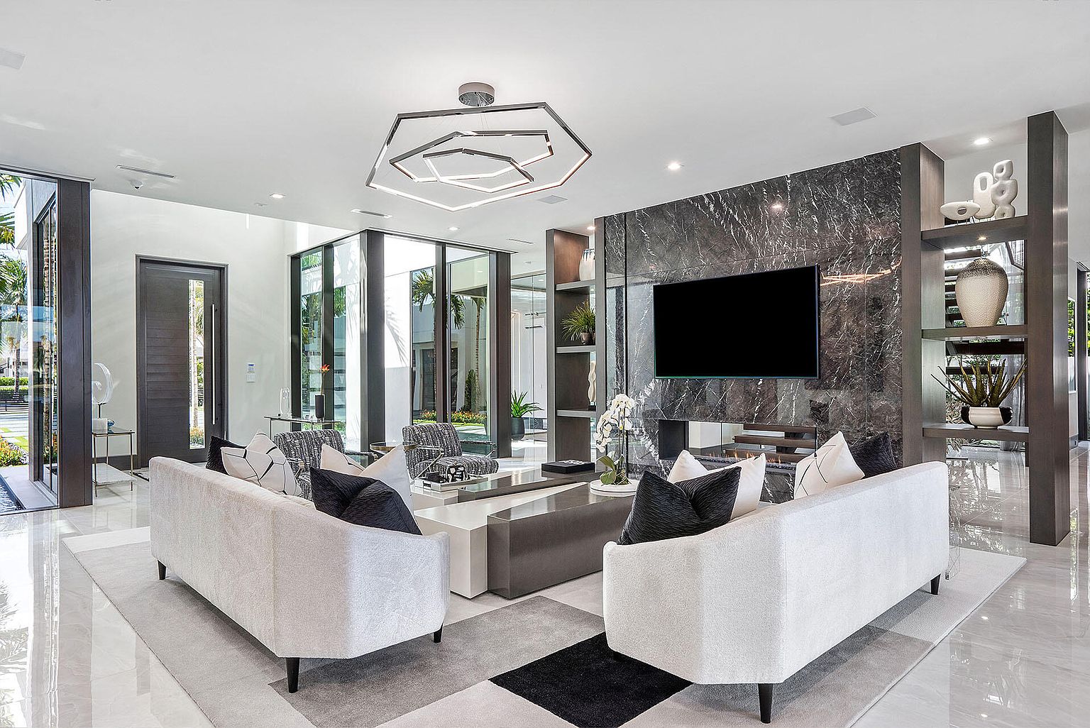This is an interior shot of a modern living room featuring two white sofas with black pillows facing each other, separated by a gray coffee table. A large marble wall with a built-in shelving unit and a flat-screen TV serves as a focal point. The room is well-lit with a unique geometric chandelier and natural light, creating a luxurious and inviting atmosphere.