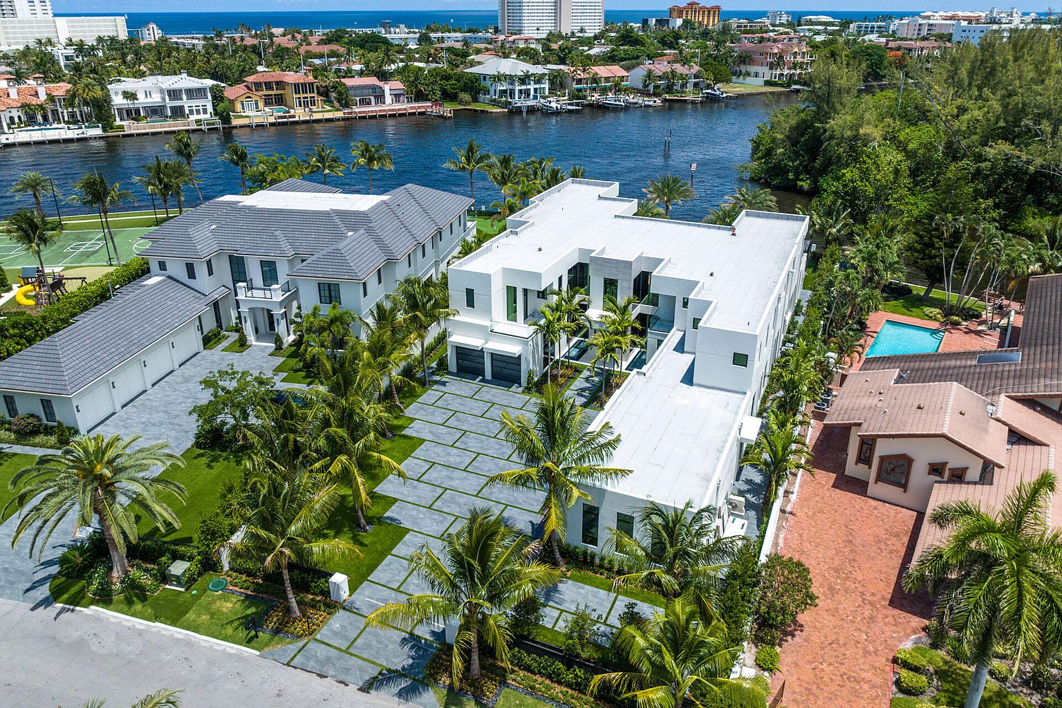 This aerial shot showcases two luxurious waterfront homes with modern architectural designs. The property features meticulously landscaped grounds with palm trees, a spacious driveway with geometric paving, and a private tennis court. The overall impression is one of high-end living and sophisticated design, set against a backdrop of serene waterways and lush greenery.