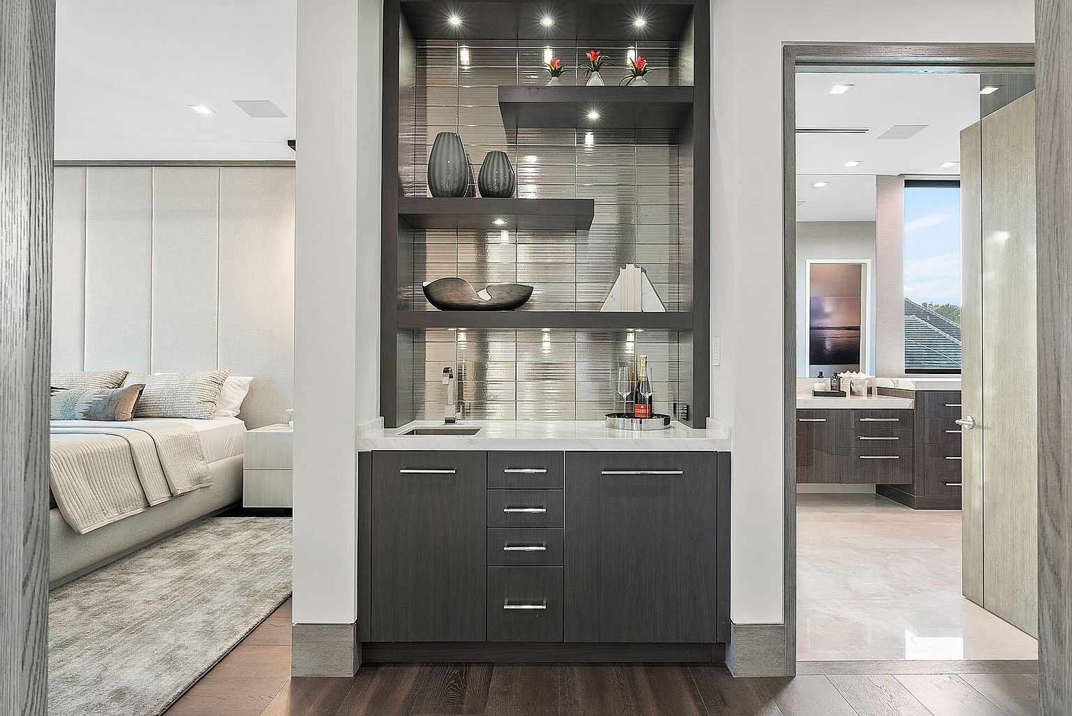 This image showcases a luxurious primary bedroom with an adjacent bathroom, connected by a stylish wet bar area. The bedroom features a padded headboard and neutral tones, while the bathroom boasts modern cabinetry and fixtures. The wet bar includes shelving with decorative items and a small sink, adding a touch of elegance and convenience.