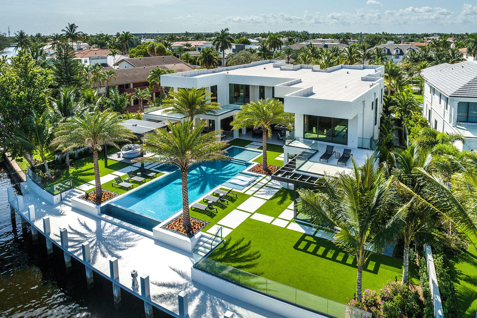 This aerial shot showcases a luxurious modern home with a stunning backyard oasis. The property features a large swimming pool with lounge chairs, manicured green spaces with geometric pavers, and mature palm trees. A private dock extends into the water, enhancing the waterfront appeal of this high-end residence.
