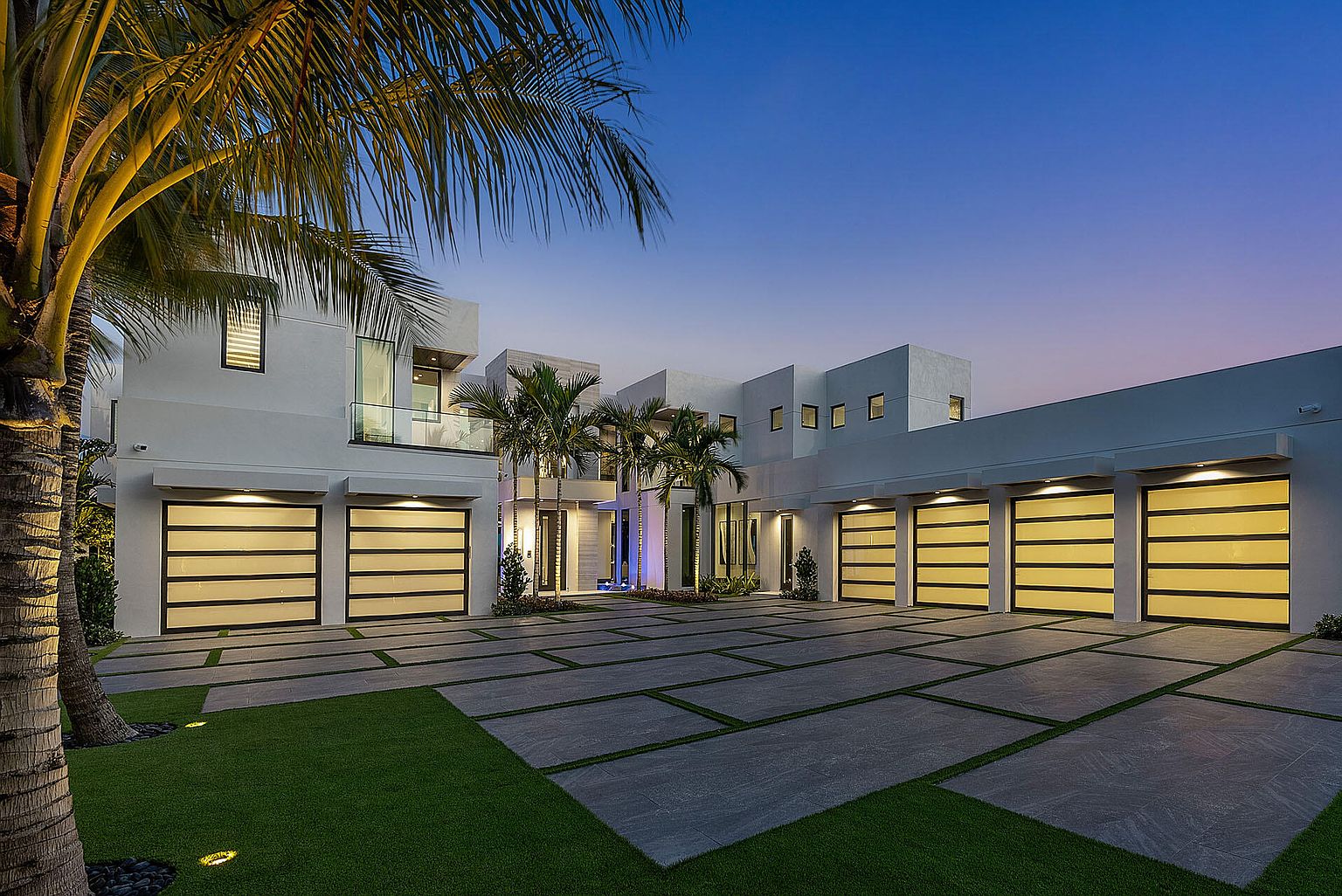 This is a front exterior view of a modern, luxurious home at dusk. The house features a multi-car garage with illuminated doors, a spacious driveway paved with geometric concrete slabs interspersed with grass, and palm trees adding a tropical touch. The architecture is contemporary with clean lines and a white facade, creating an inviting and upscale impression.