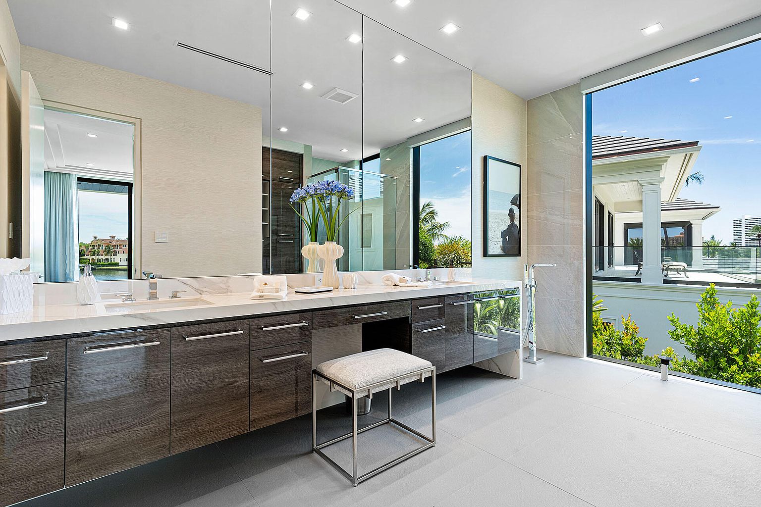 This is a luxurious primary bathroom featuring a long, dark wood vanity with a white marble countertop and dual sinks. A large mirror spans the length of the vanity, reflecting natural light from a large window that offers a view of the exterior. The bathroom is spacious and well-lit, with a modern and elegant design.