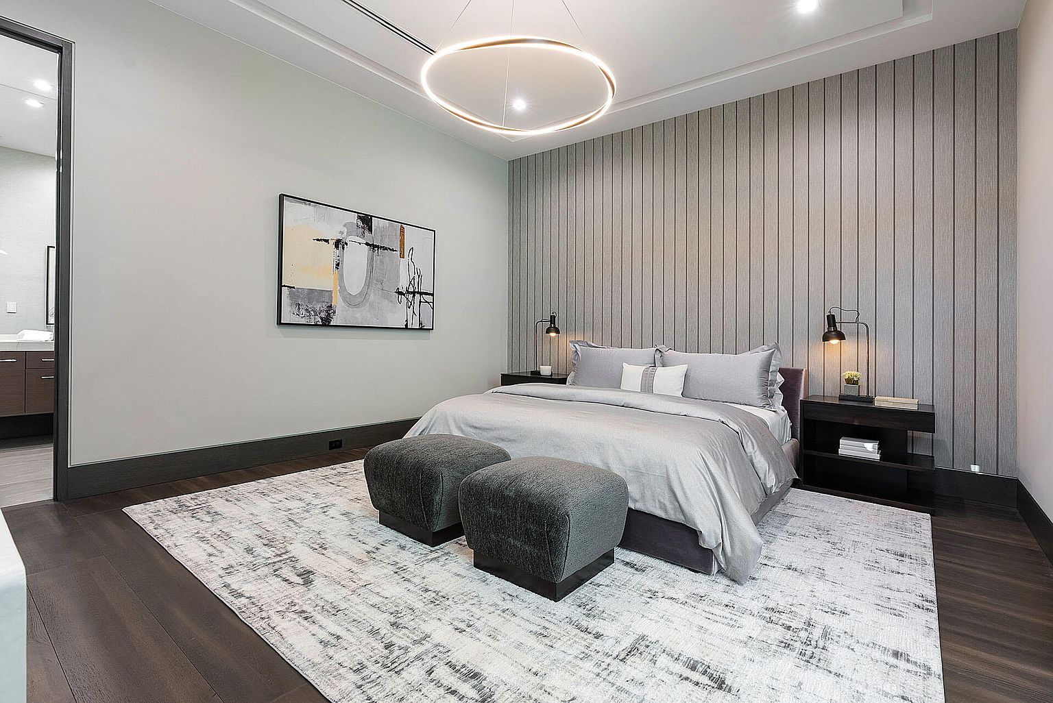 This is a primary bedroom featuring a king-size bed with gray bedding, flanked by dark wood nightstands and modern lamps. The room is decorated with a large abstract painting and a unique circular chandelier. The flooring is dark wood, covered by a light-colored area rug, and two gray ottomans are placed at the foot of the bed.